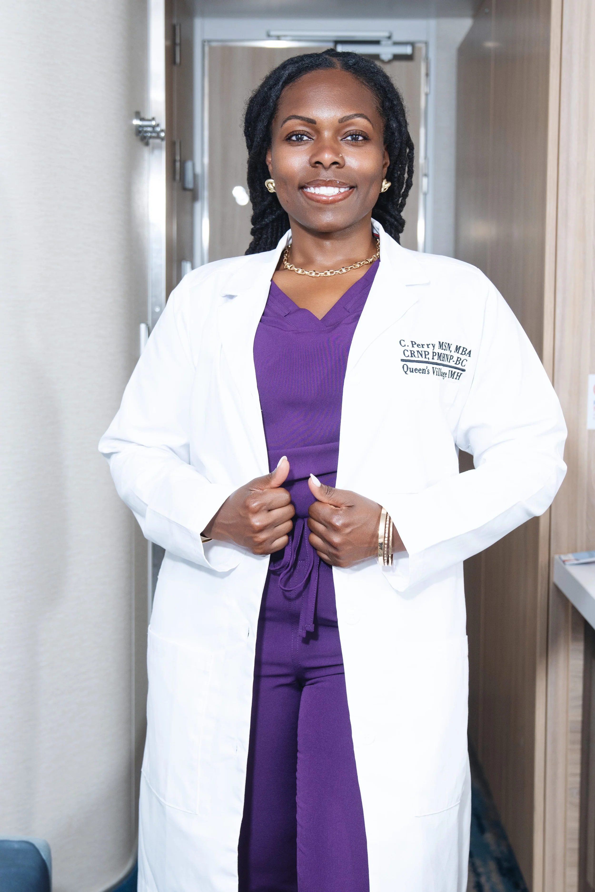 A confident female healthcare professional standing in a medical facility, wearing a white coat with her name and credentials, purple scrubs, gold jewelry, and smiling.