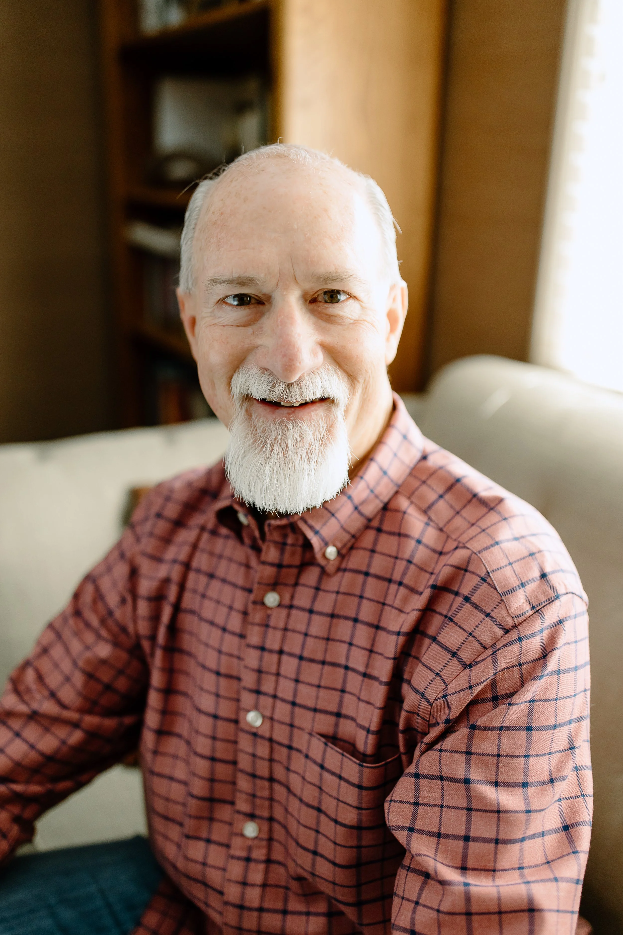 A smiling elderly man with a white beard and mustache wearing a checkered shirt and sitting indoors with a bookshelf in the background.