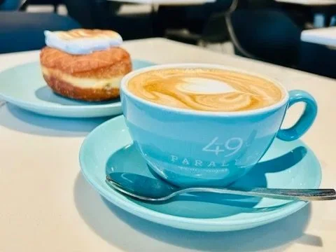 A blue cup of coffee with latte art on top, on a matching saucer with a spoon, and a dessert pastry on a blue plate in the background.