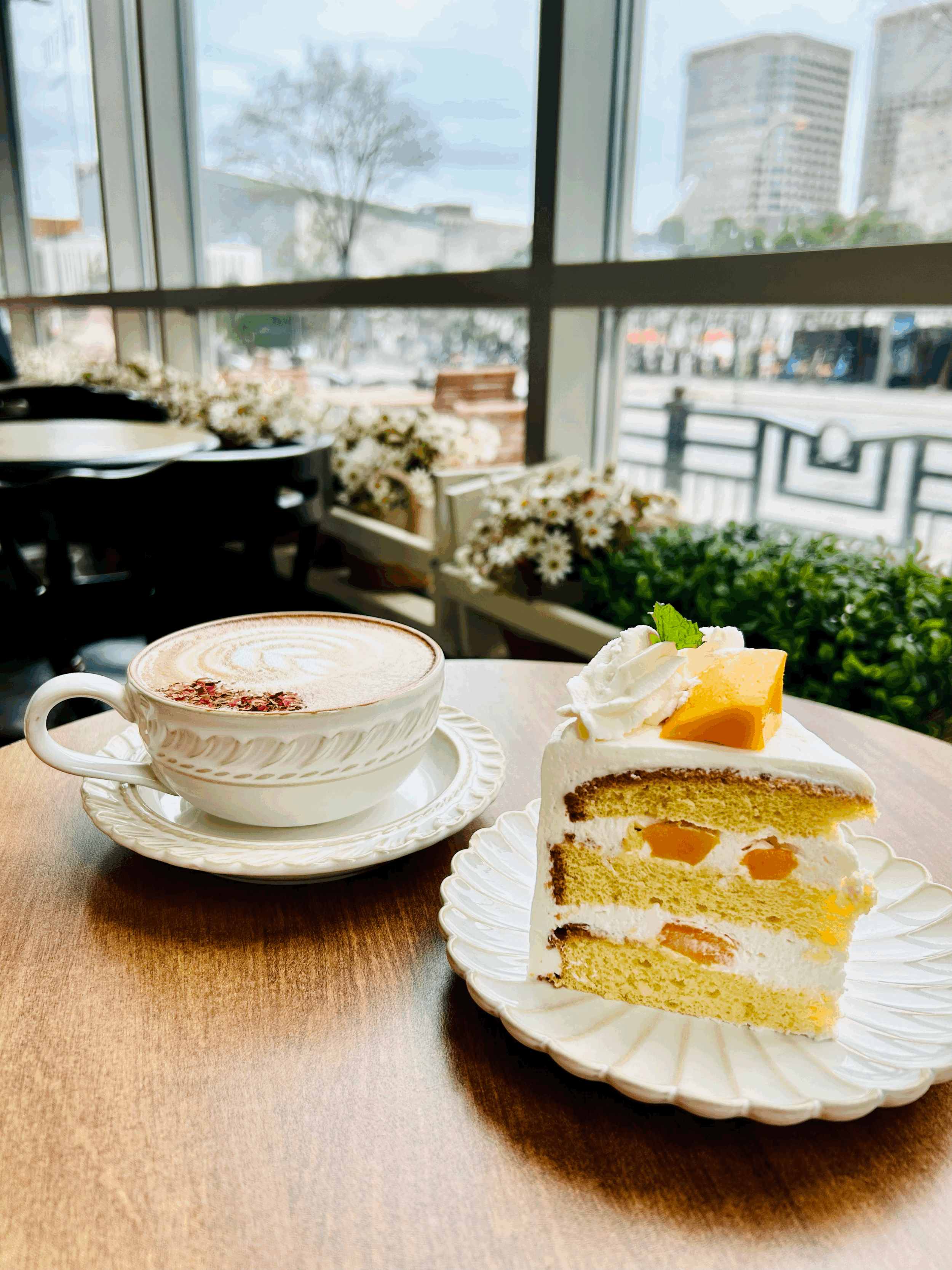 A cup of coffee with foam and flower petals and a slice of layered fruit cake with cream, fruit filling, and a topping with whipped cream and a mint leaf on a white plate on a wooden table in a cafe, with large windows and an urban view outside.