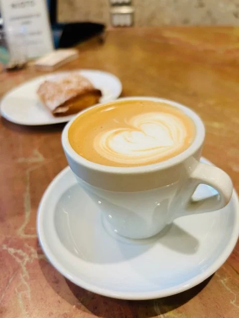 White ceramic cup of latte with heart-shaped latte art on top, placed on a matching saucer on a wooden table, with a plate of croissant in the background.