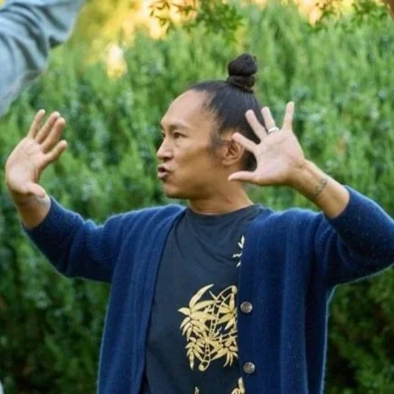 Jonathan Nobleza wearing a dark blue cardigan over a black shirt with a gold design, standing outdoors with green foliage in the background, making expressive hand gestures.