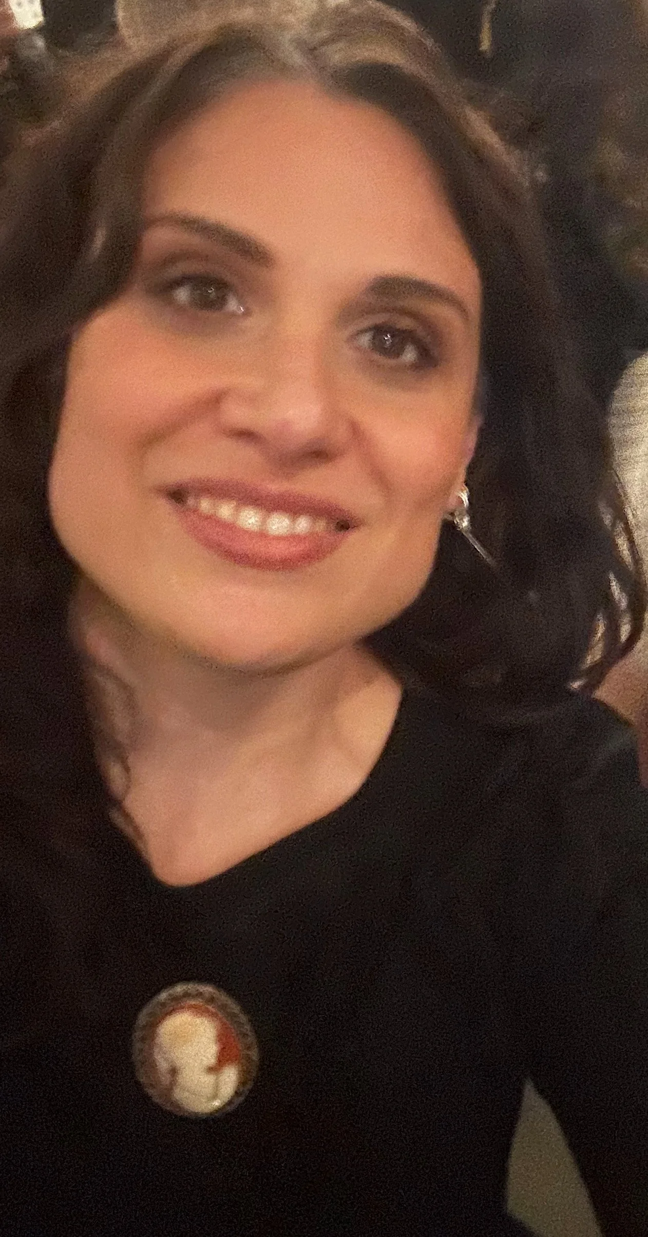 A woman with dark, wavy hair and light skin smiling at the camera. She is wearing earrings and a black top with a button featuring a WWII soldier's face.