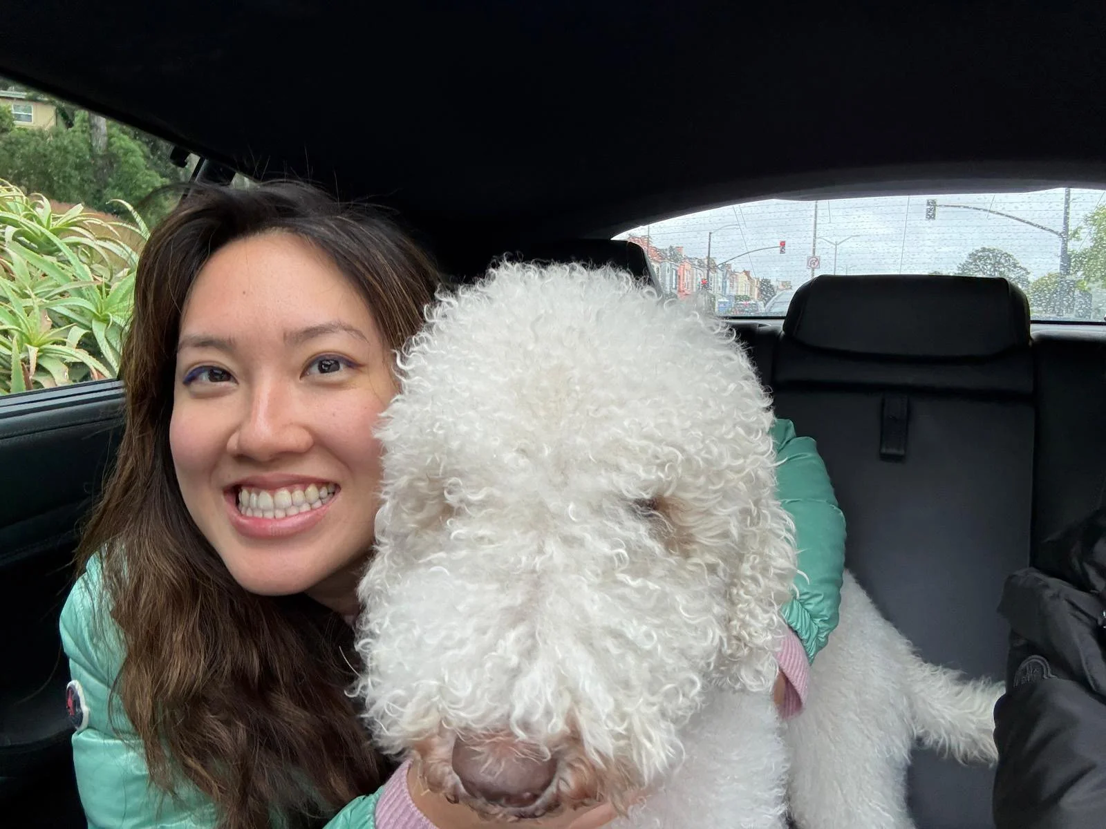 A woman smiling in a car with a white curly-haired dog. The woman has long brown hair, is wearing a teal jacket, and is sitting next to the dog. The background shows a rainy day through the car window.