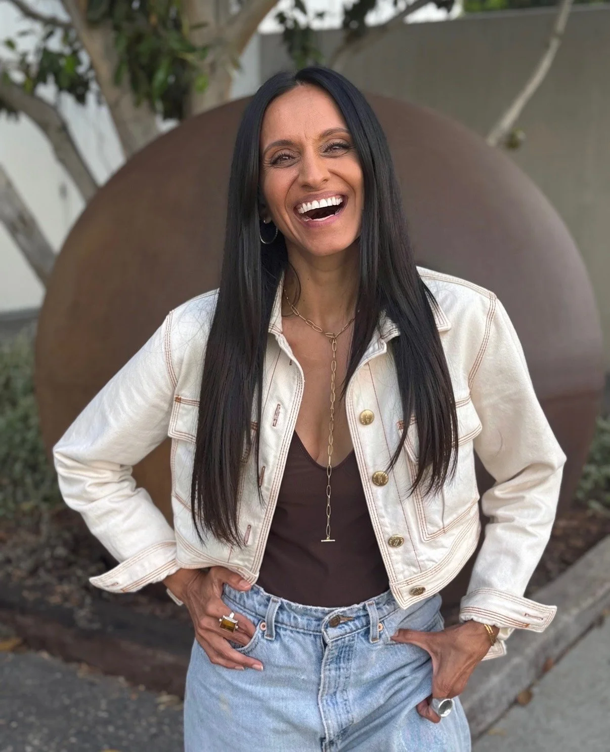 Monisha Chandanani laughing, wearing a white denim jacket over a black top, gold jewelry, and light blue jeans, standing outdoors in front of a large round sculpture and trees.