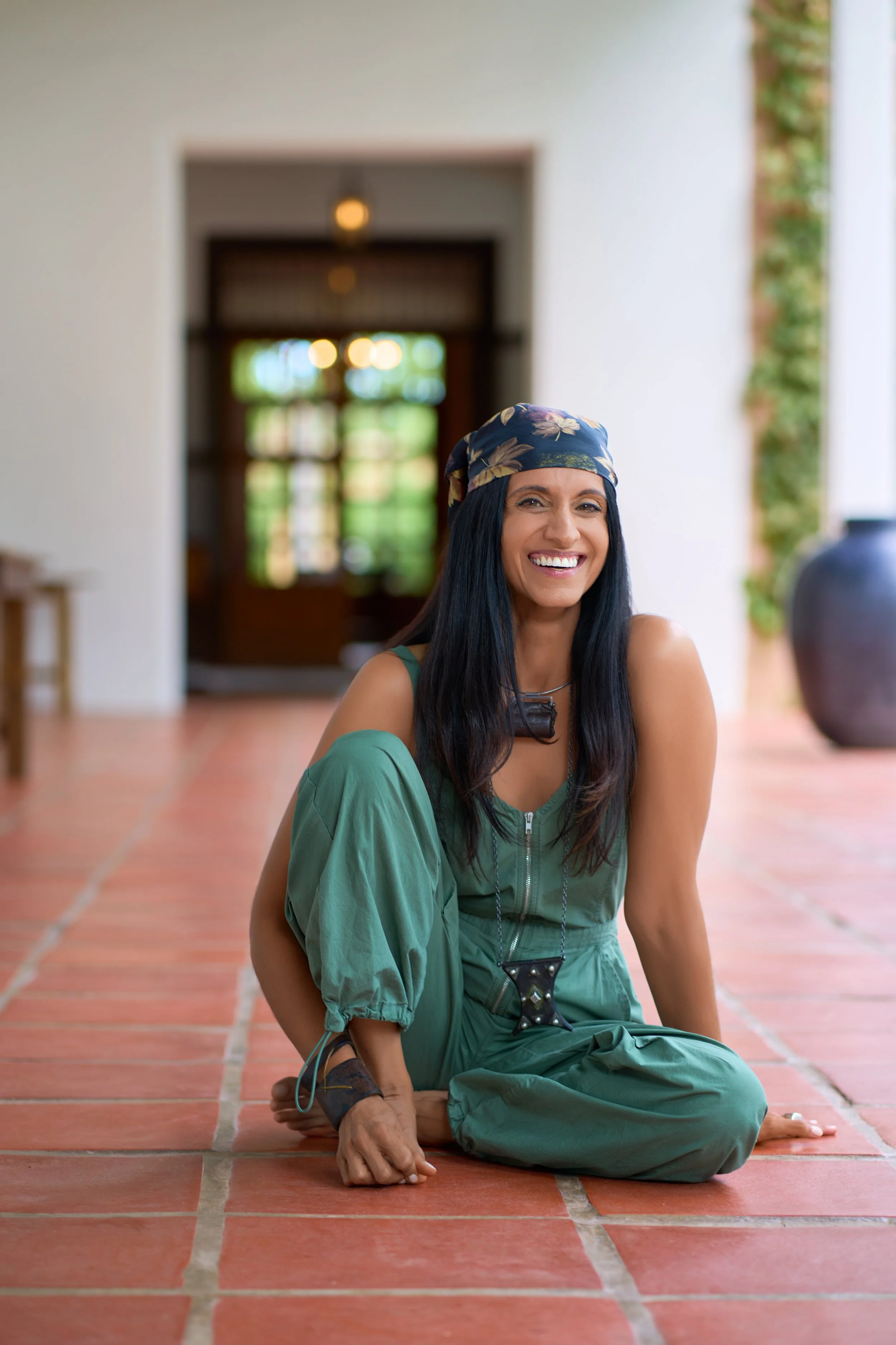 Monisha Chandanani wearing a blue floral headscarf and green jumpsuit, smiling while sitting on a terracotta tile floor indoors, with a blurred background of a room with trees outside and large pottery vase.