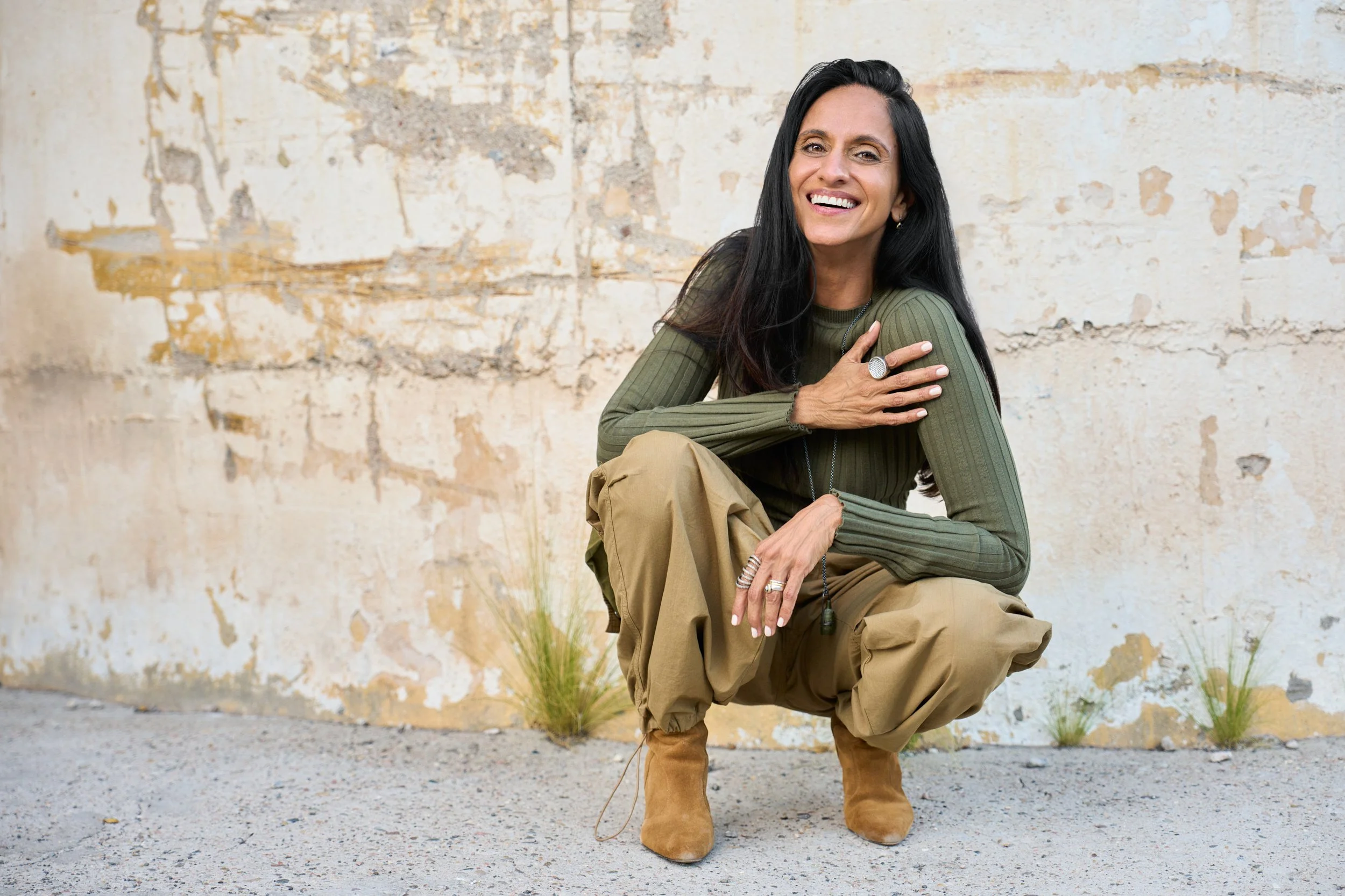 Monisha Chandanani wearing an olive green shirt and tan pants, squatting outdoors against a weathered wall, smiling.