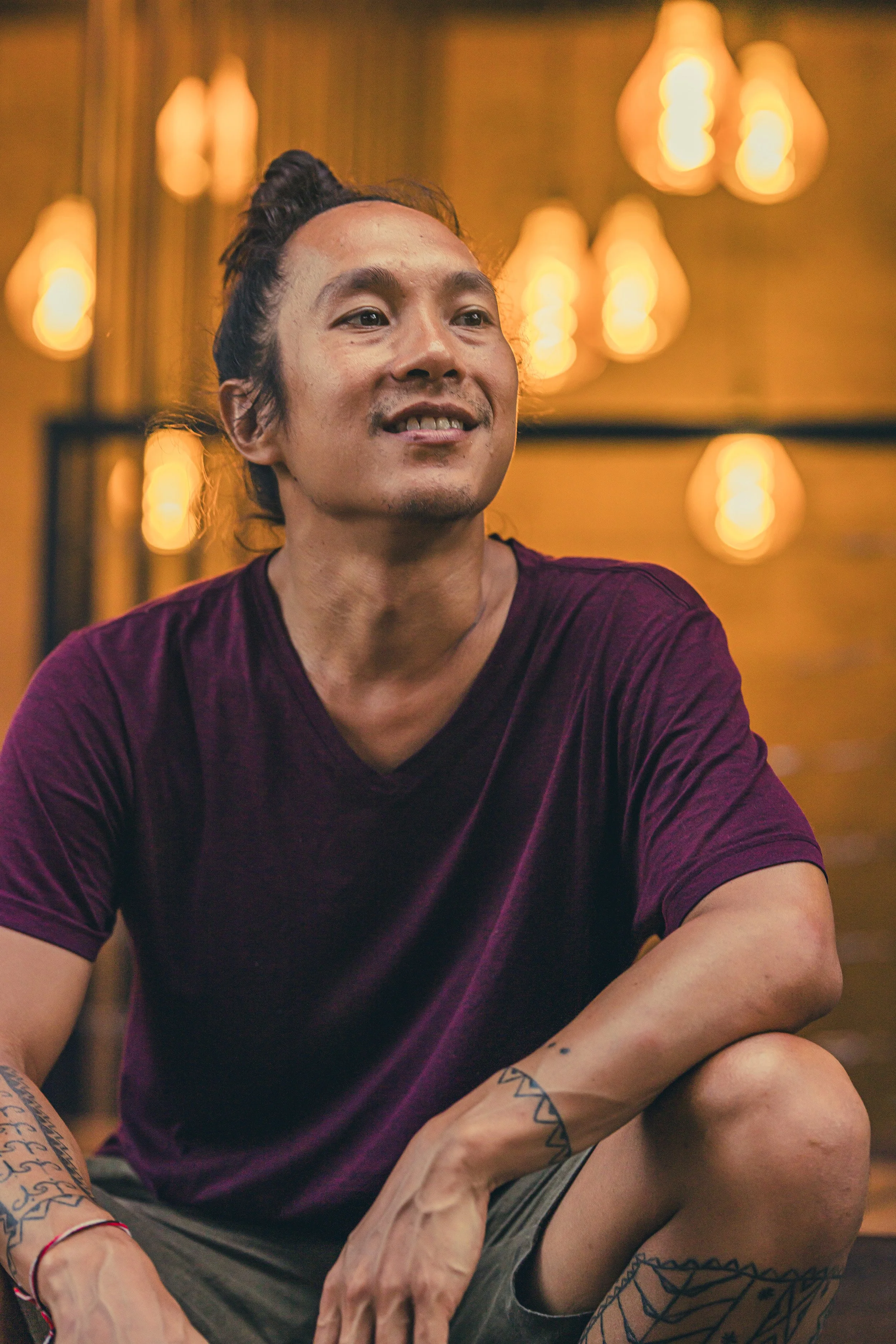 Jonathan Nobleza wearing a maroon shirt and gray shorts, sitting with one knee up, in a warmly lit setting with hanging light bulbs in the background.