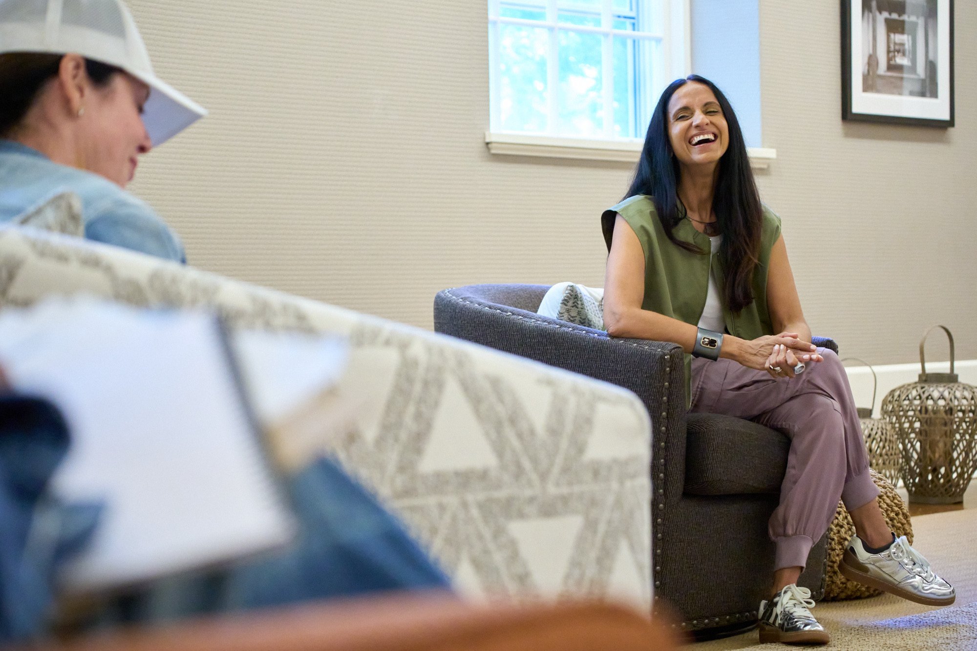 Monisha Chandanani laughing while sitting in a chair in a room with students, sunlight coming through a window.