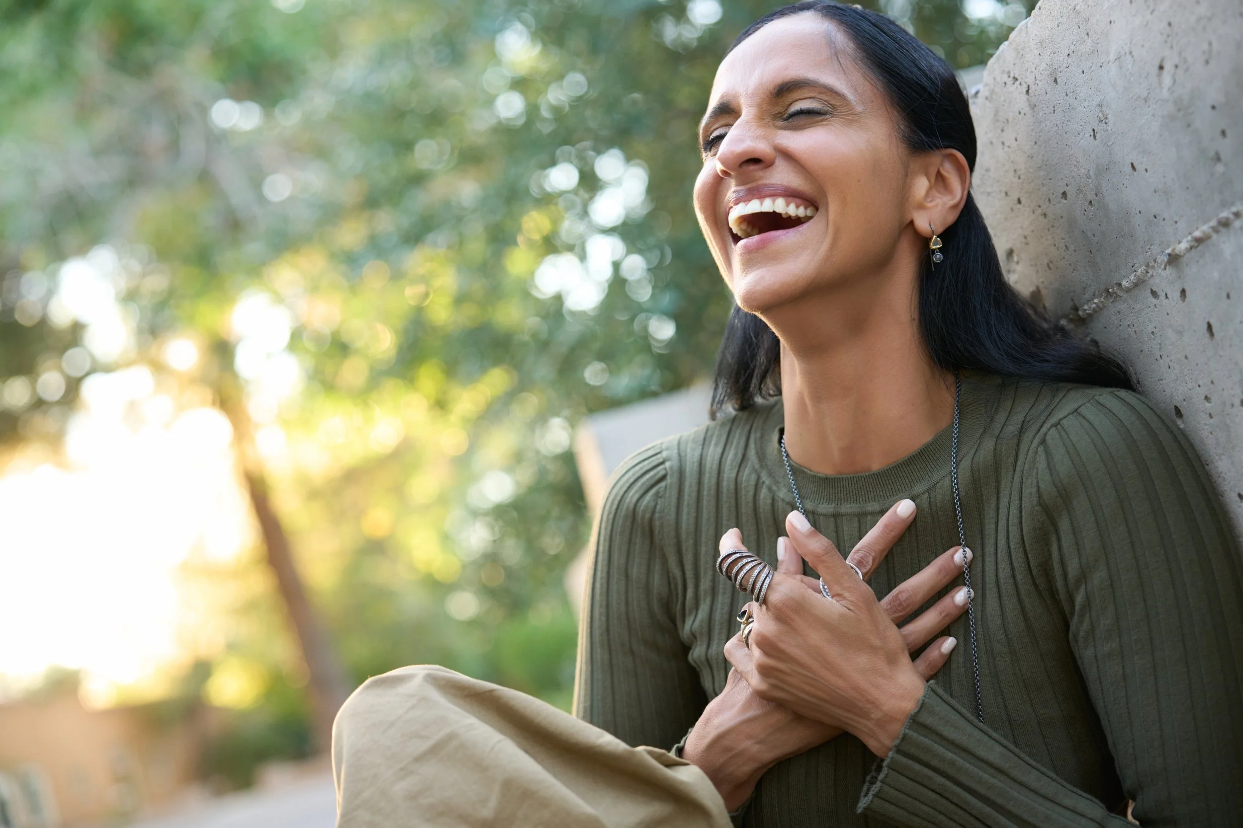 Monisha Chandanani laughing outdoors, leaning against a concrete wall, with her eyes closed and her hand over her chest, wearing rings, earrings, and a green long-sleeve shirt, with blurred greenery and sunlight in the background.