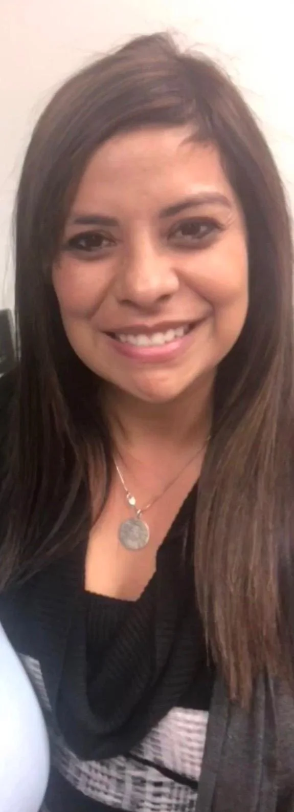 Close-up of a smiling woman with long brown hair wearing a black top and a silver necklace with a round pendant.