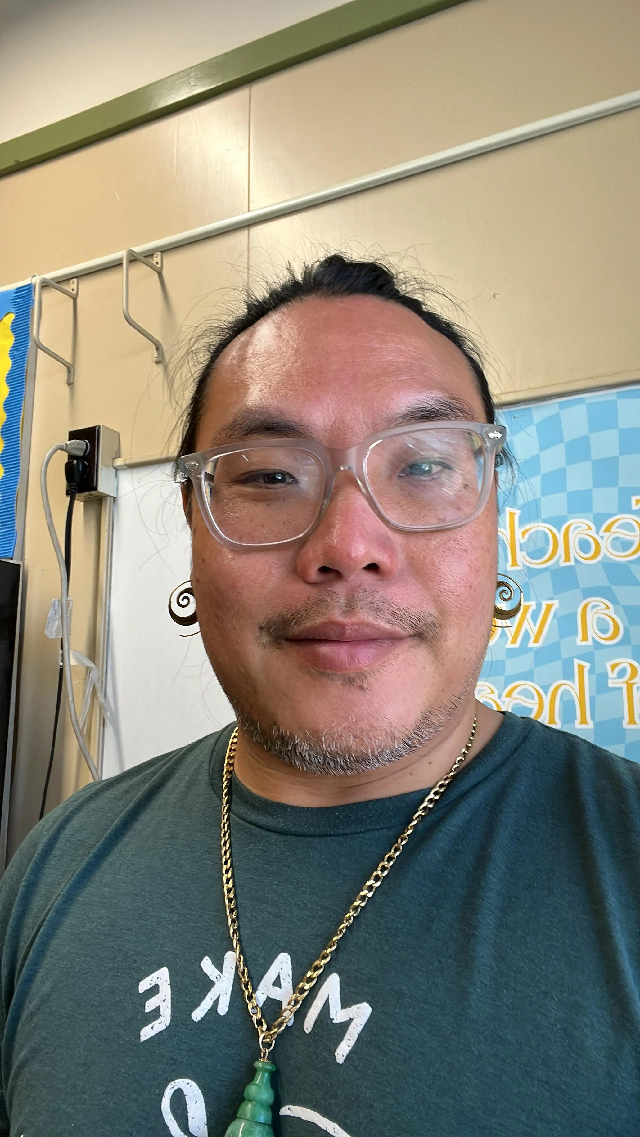 A close-up of a man with glasses, a goatee, wearing a green shirt, gold chain with green pendant, and earrings, in an indoor setting with a blue patterned background and beige and green walls.