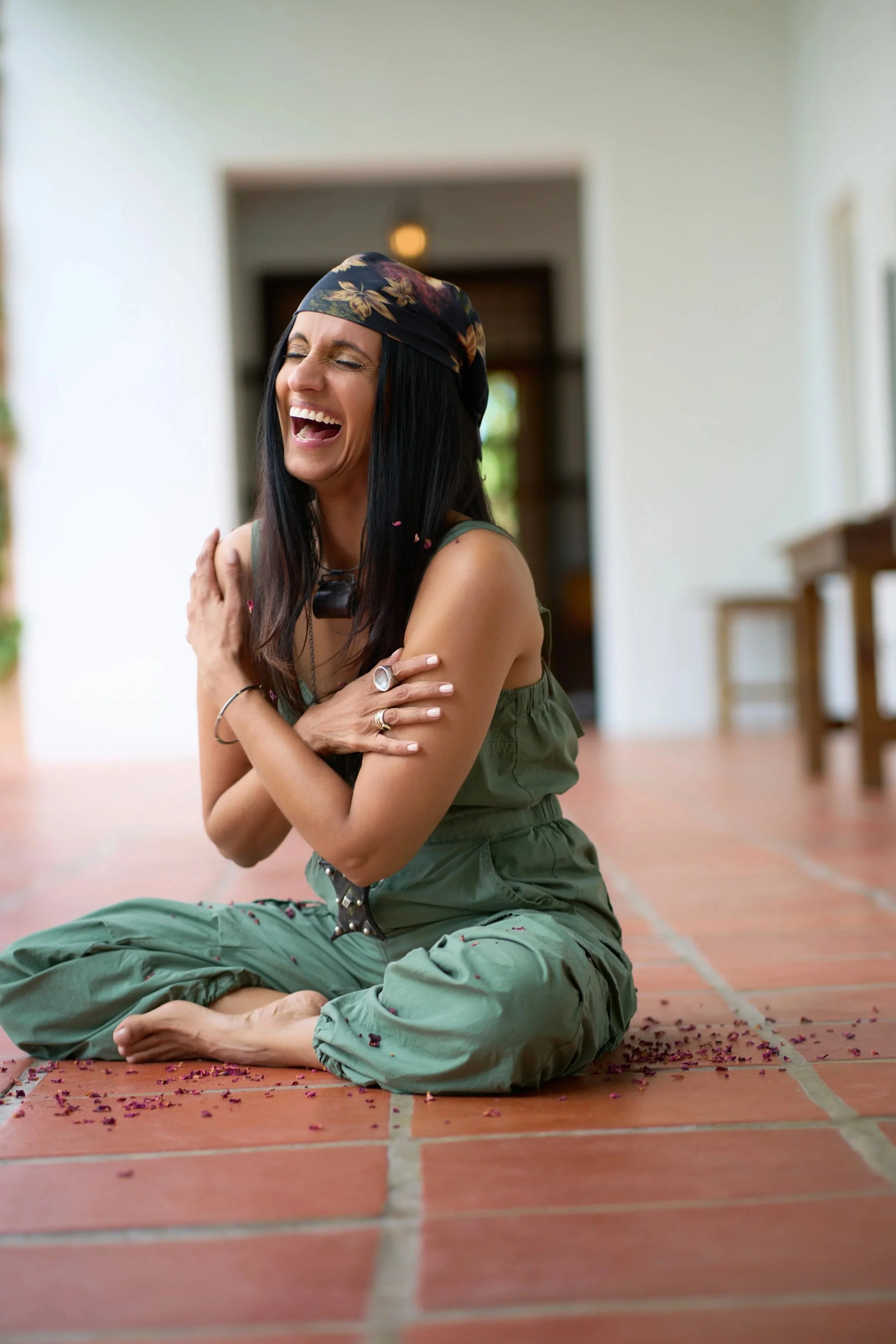 Monisha Chandanani wearing a floral headscarf and green jumpsuit, sitting cross-legged on a terracotta-tiled floor, laughing with her eyes closed, surrounded by scattered purple flower petals in a bright, airy room.