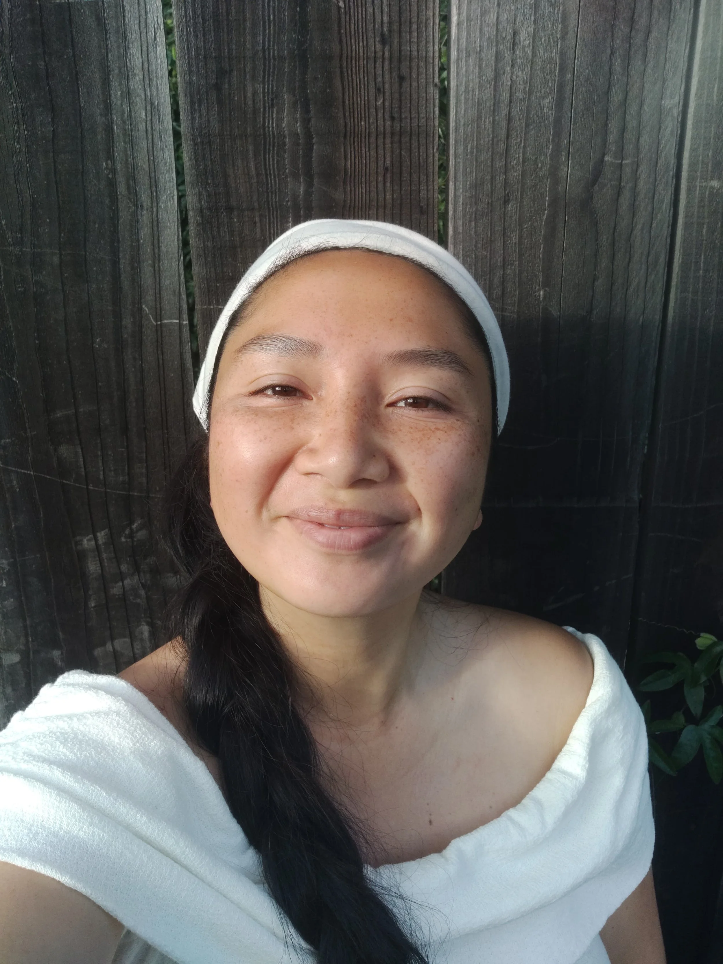 A woman with long dark hair and brown eyes smiling while taking a selfie outdoors. She is wearing a white off-the-shoulder top with a textured fabric. Behind her is a black wooden fence, and some greenery is visible through a gap in the fence.