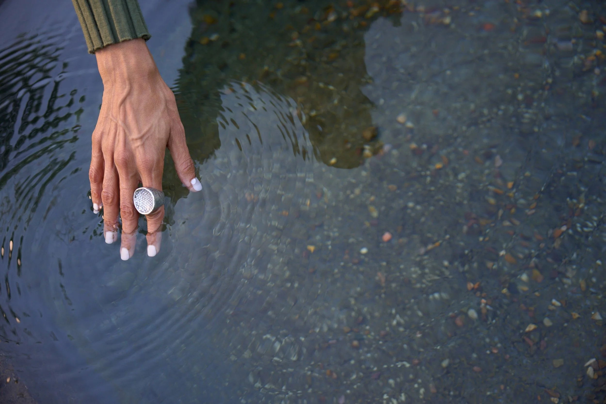 A hand reaches into clear water, with a large silver ring on the middle finger and a ribbed green sleeve partially visible. The water is shallow and reveals small pebbles at the bottom.