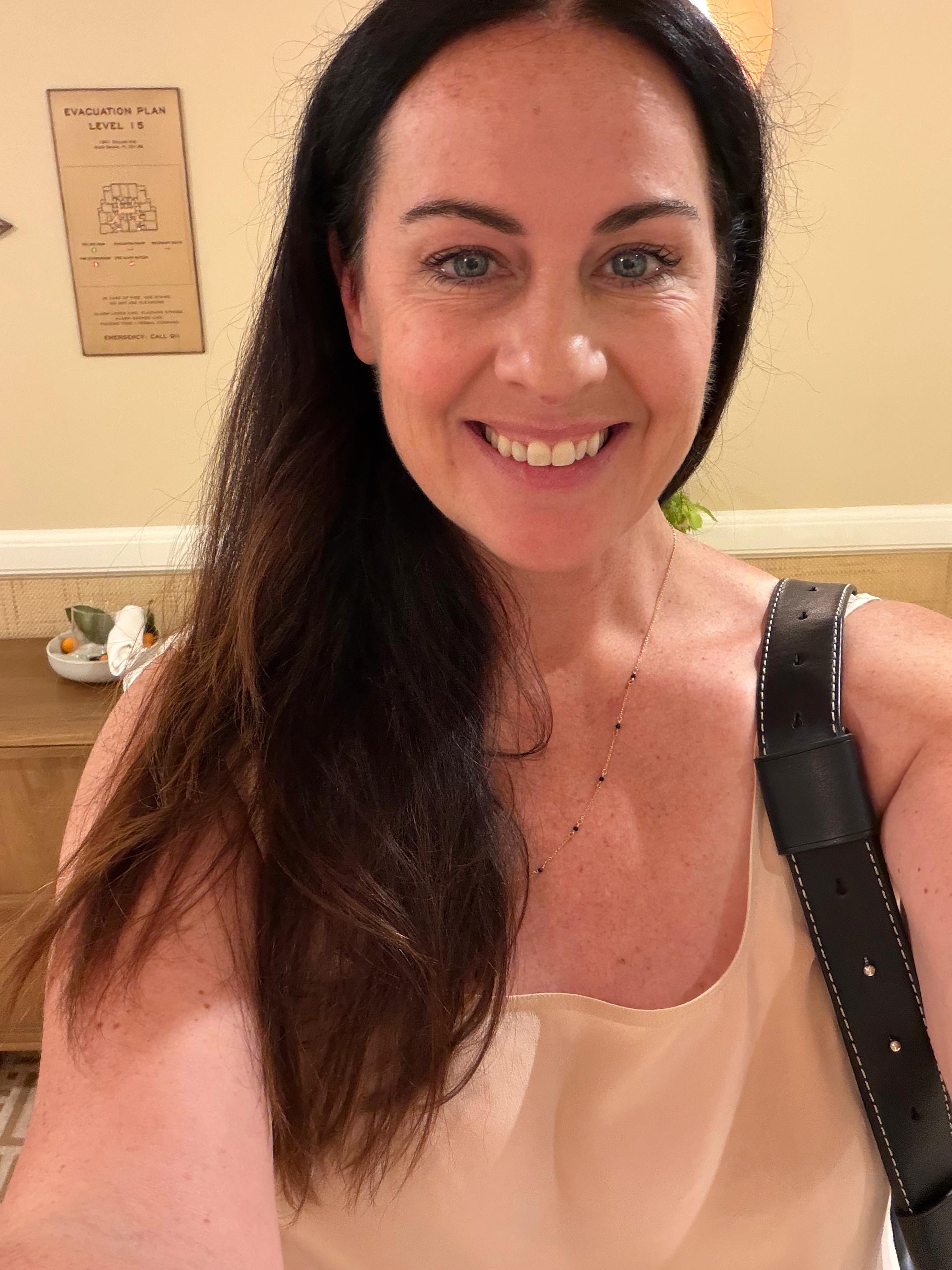 A woman with long dark hair and blue eyes smiling, wearing a light-colored top and a gold necklace, with a black strap over her shoulder, standing indoors with beige walls and a wooden table in the background.