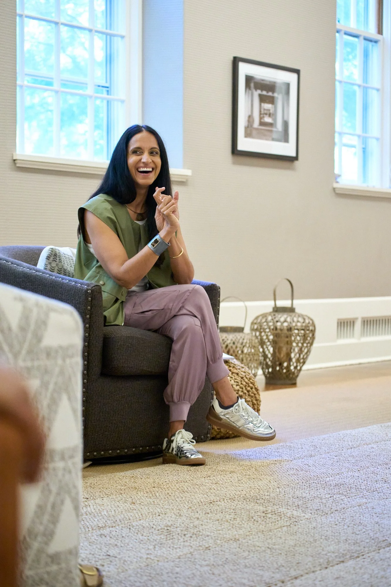 Monisha Chandanani smiling and sitting on a dark-colored sofa in a well-lit room with beige carpet, off-white walls, and large windows. She is leading a group session.
