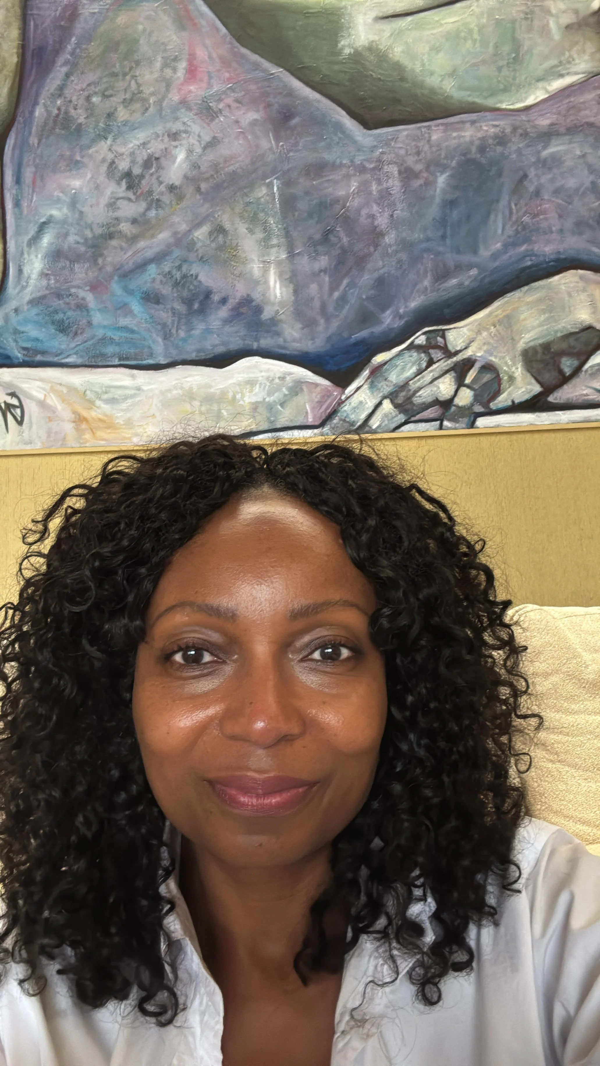 A woman with curly black hair taking a selfie in front of an abstract painting with various shades of gray, purple, green, and light colors, hanging on a yellow wall.