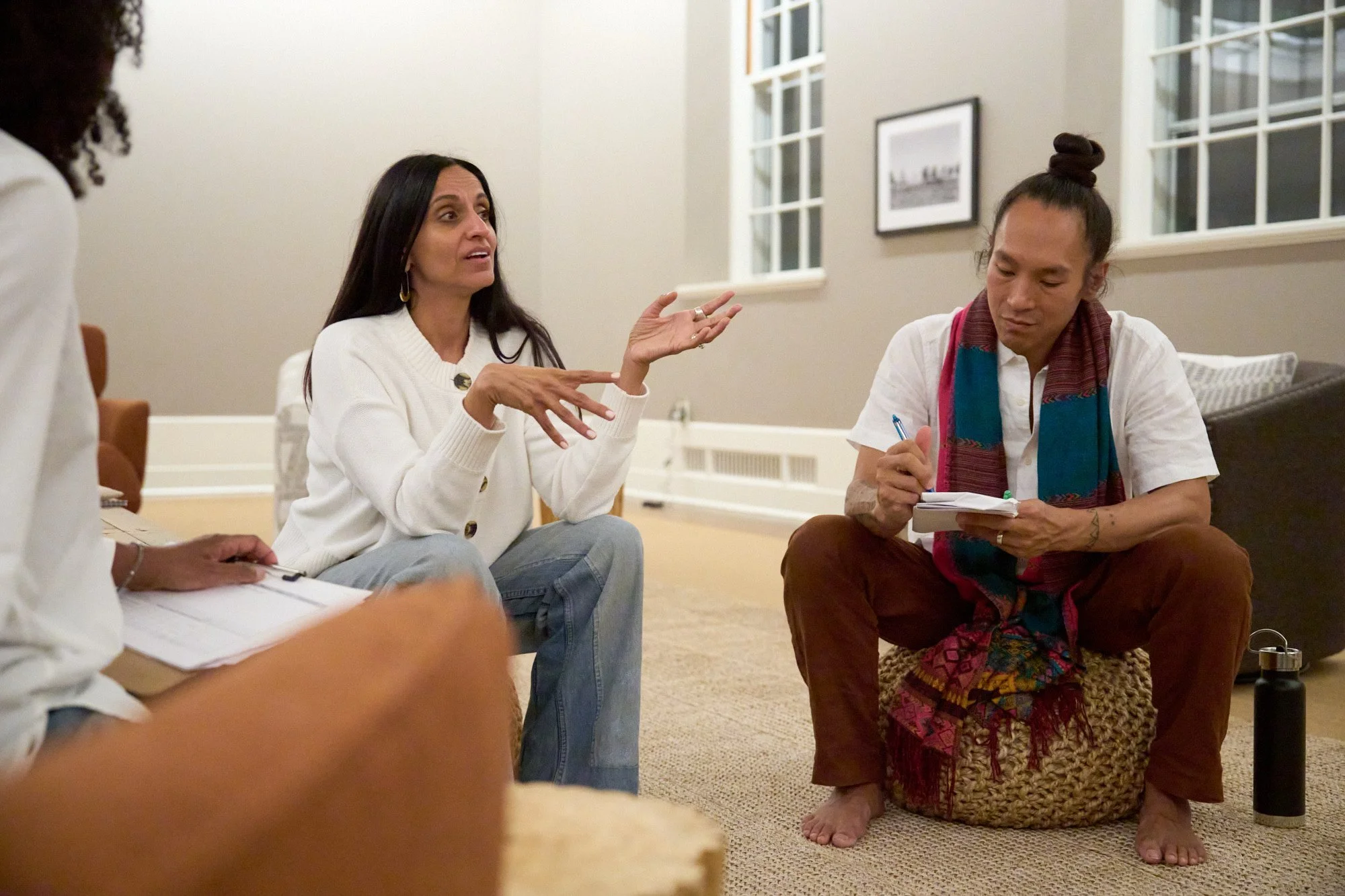 A group of diverse individuals sitting on the floor in a room, engaged in a discussion. Monisha Chandanani is speaking, gesturing with her hands. Jonathan Nobleza is taking notes on a notepad.