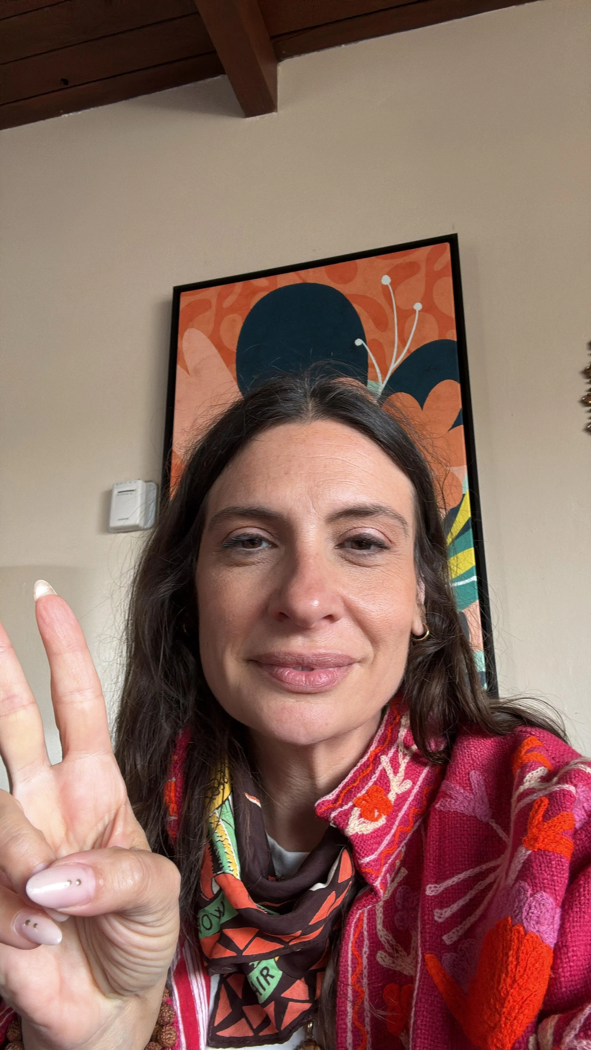 A woman taking a selfie indoors with a colorful abstract painting in the background. She is making a peace sign with her left hand and has dark hair, light makeup, and is wearing a vibrant red patterned jacket and a multicolored scarf.