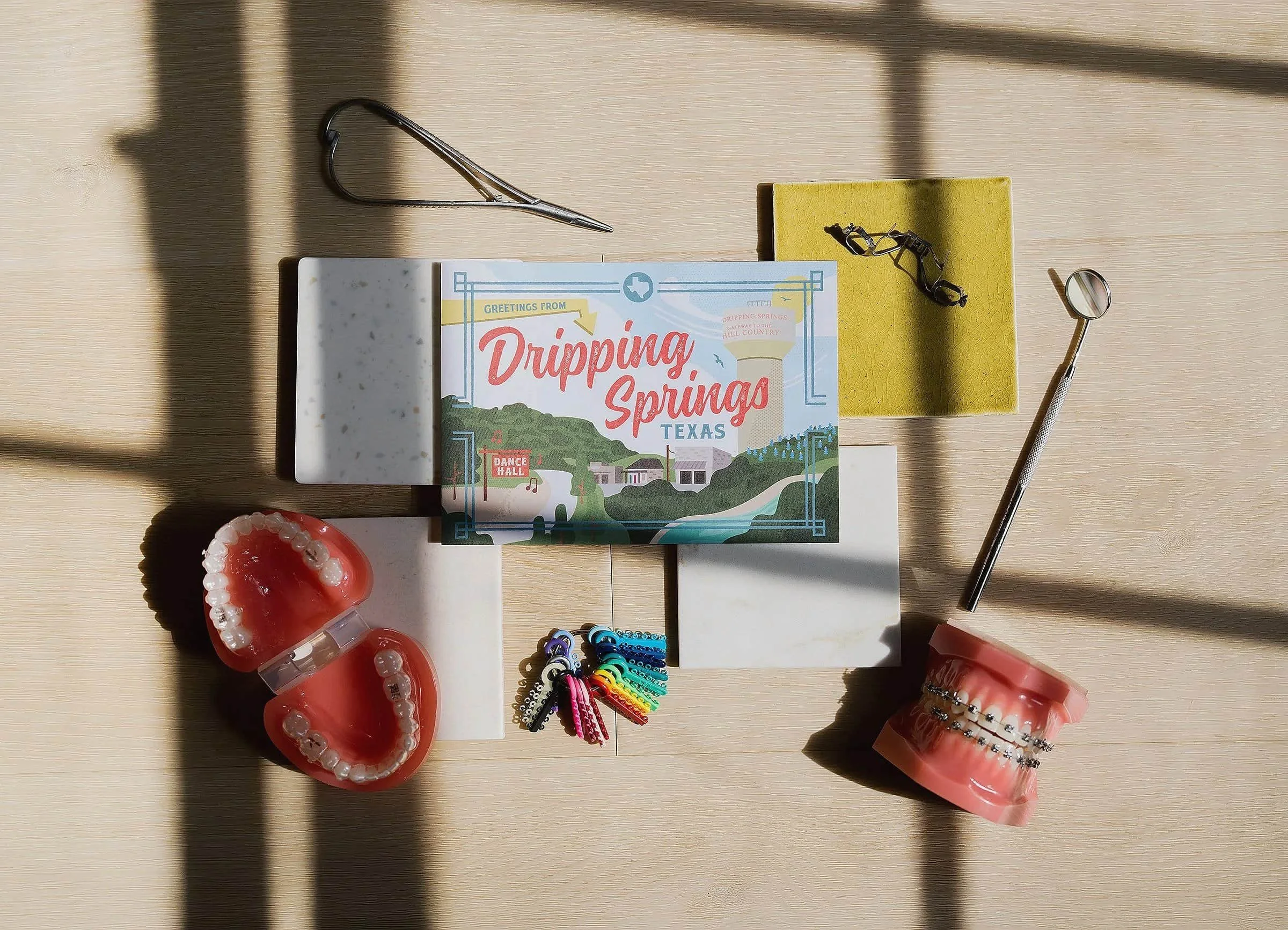 Gil Orthodontics brand display featuring a Dripping Springs Texas postcard, clear aligner trays, traditional braces models, and an orthodontic color wheel on top of interior design material swatches.