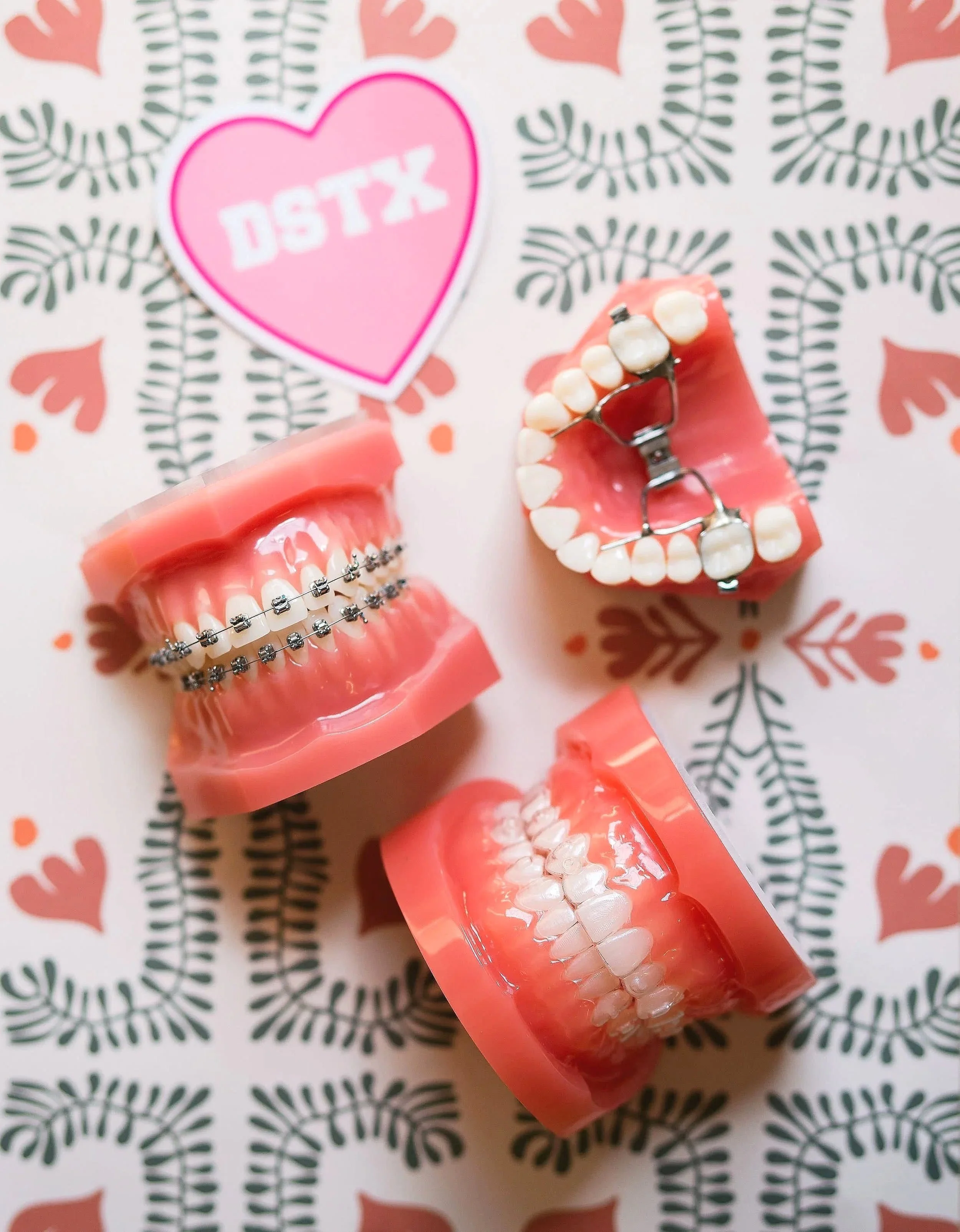 Dental models displaying traditional metal braces, clear Angel aligners and a palatal expander next to a pink DSTX heart sticker representing Dripping Springs.