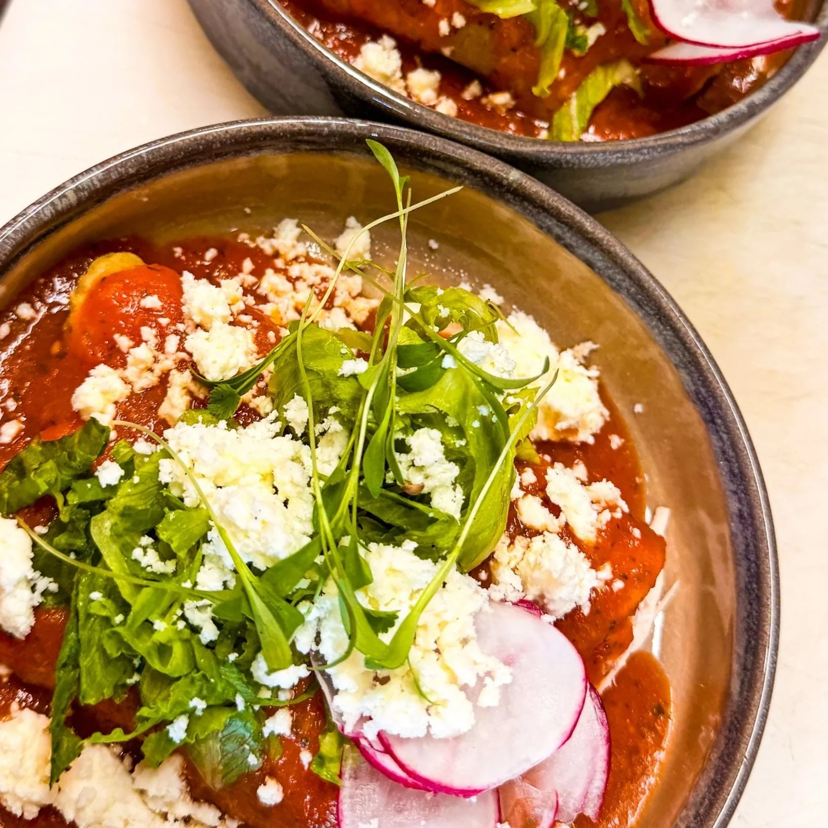 Close-up of a bowl containing Mexican soup topped with fresh greens, radish slices, crumbled cheese, and shredded chicken.