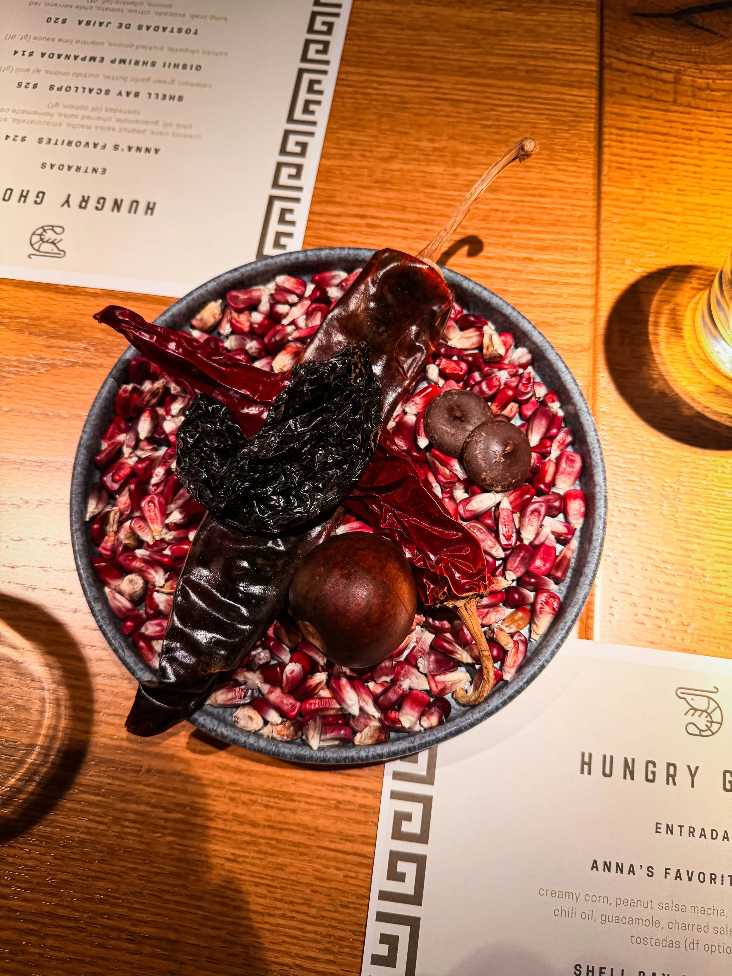 A plate of seasoned popcorn with dried chili peppers and two dried dark red chili peppers on top, placed on a wooden table with a partially visible menu in the background.