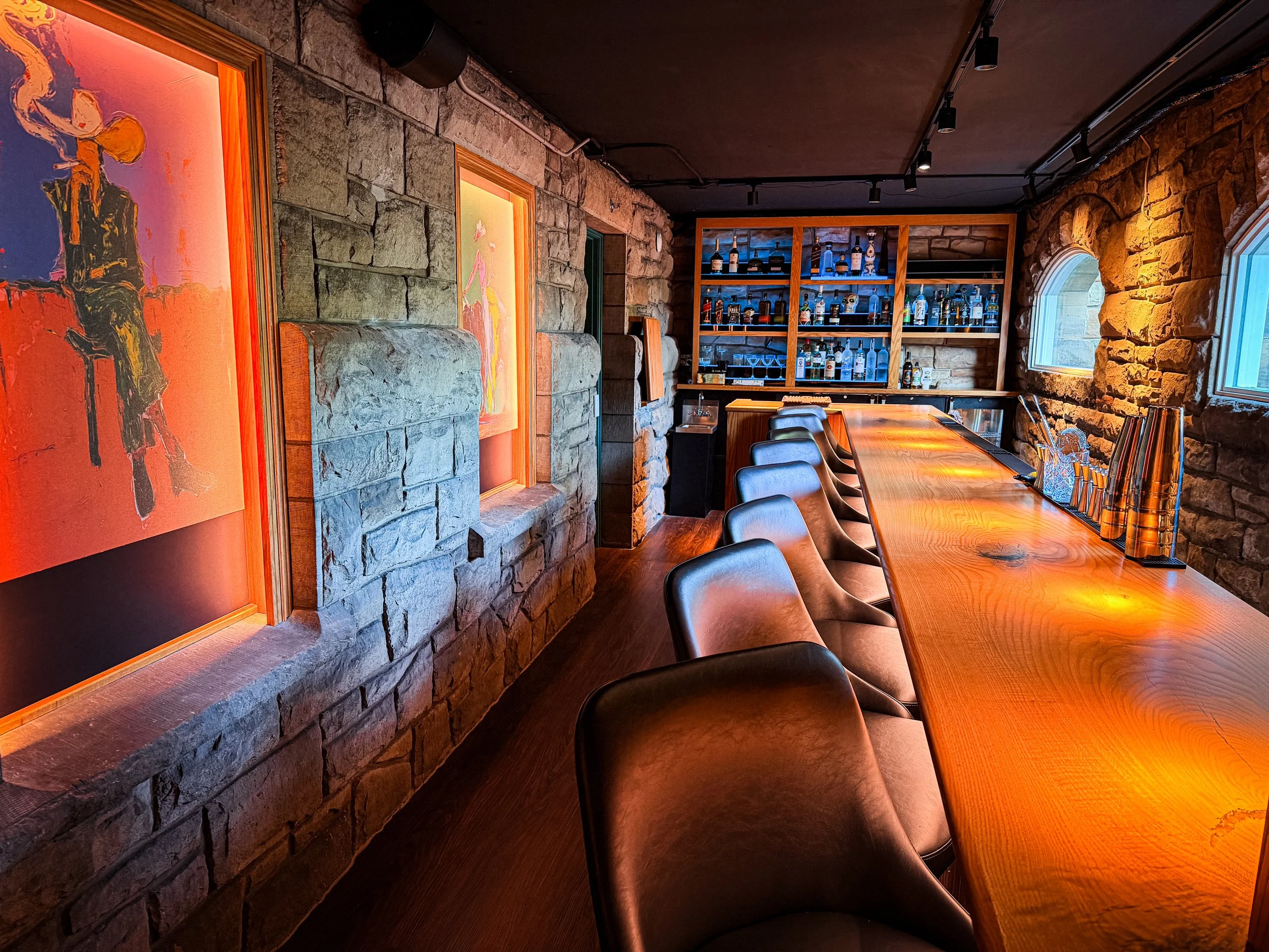 Interior of a bar or restaurant with a long wooden table, black chairs, stone walls, colorful paintings, a bar shelf with bottles, and small windows letting in natural light.