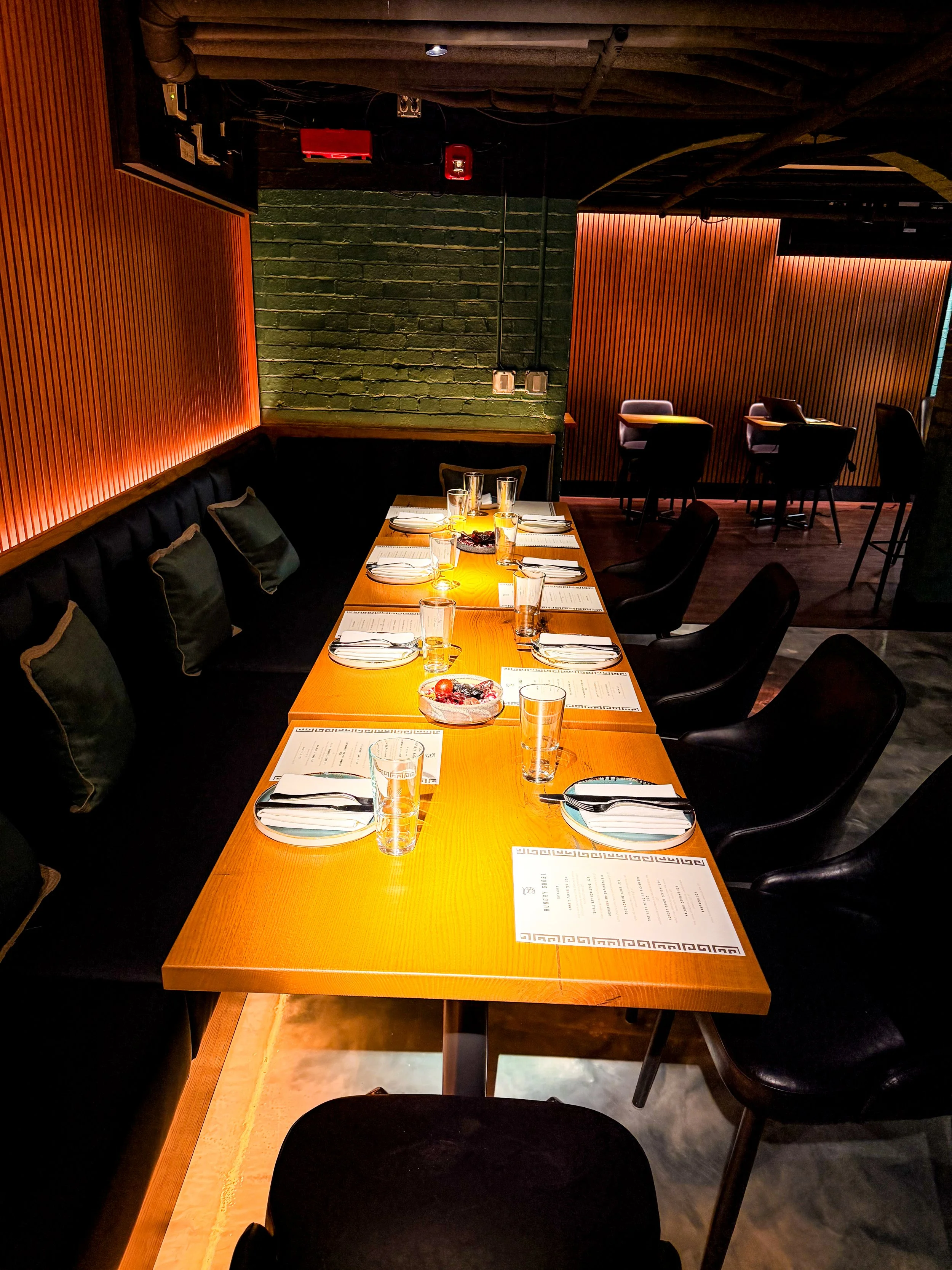 A long wooden dining table set with plates, glasses, and silverware, with black chairs around it in a dimly lit restaurant featuring green brick walls and wooden paneled decor.