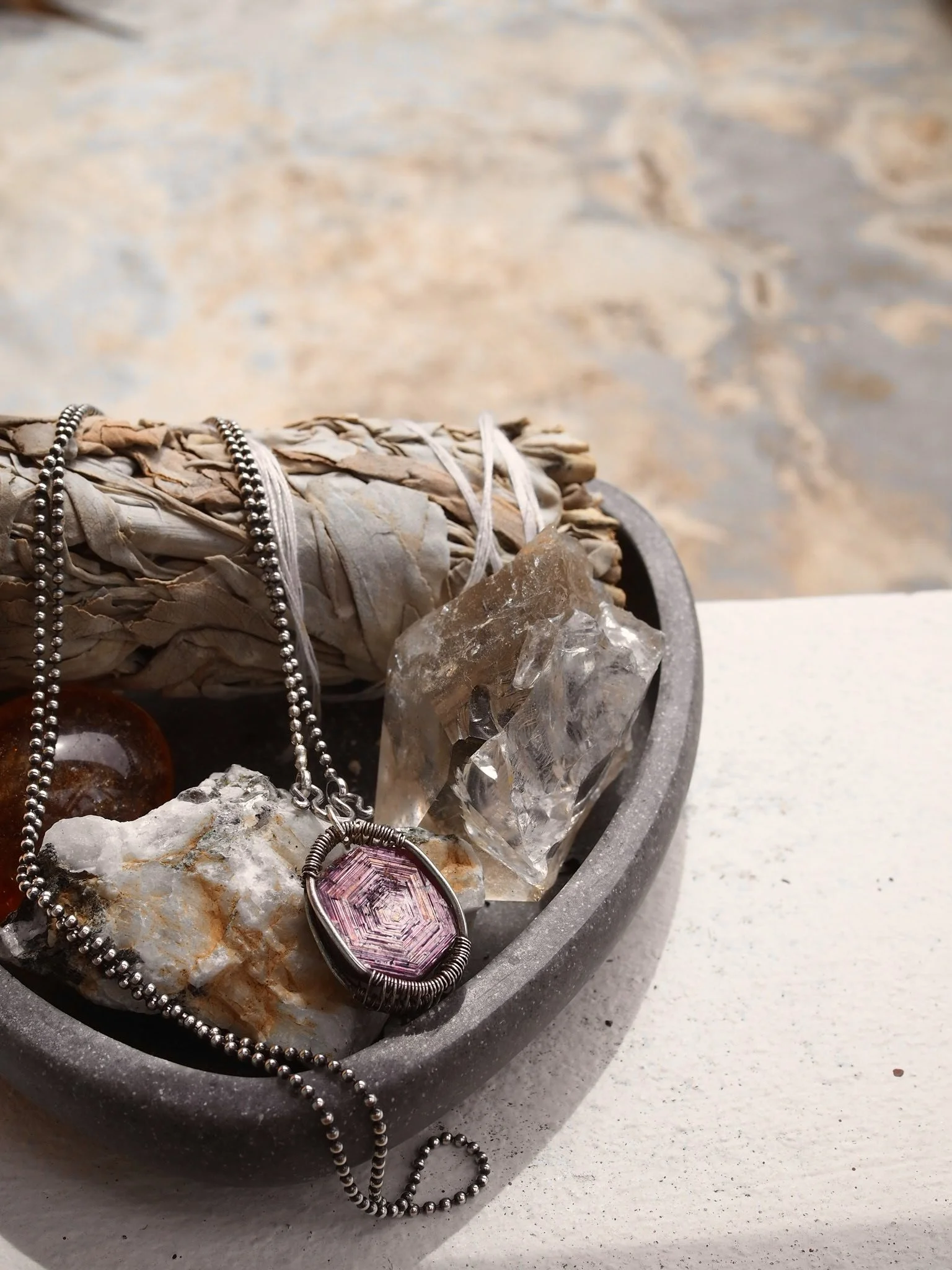 A close-up of a jewelry collection including a pink gemstone pendant, a silver bead necklace, and a crystal, placed on a bed of rocks inside a gray dish.