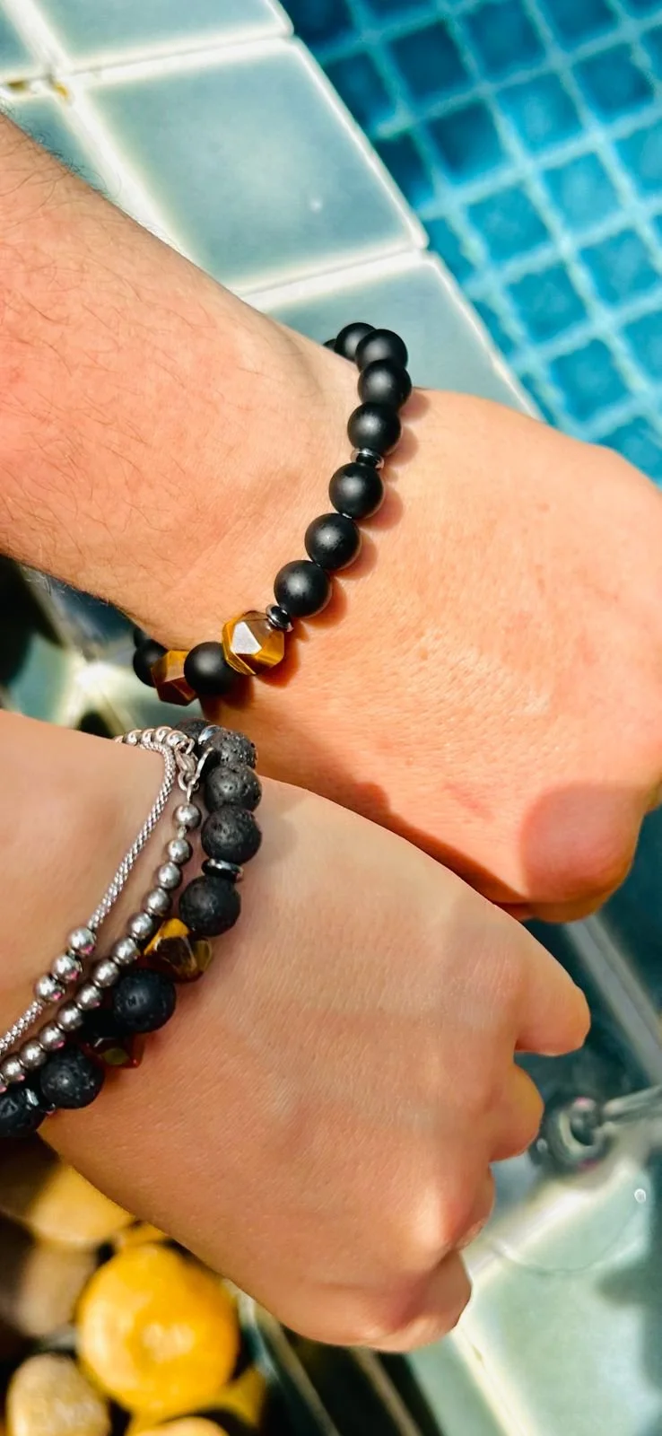 Close-up of two people holding hands over a sink with water, wearing matching black and gold beaded bracelets.