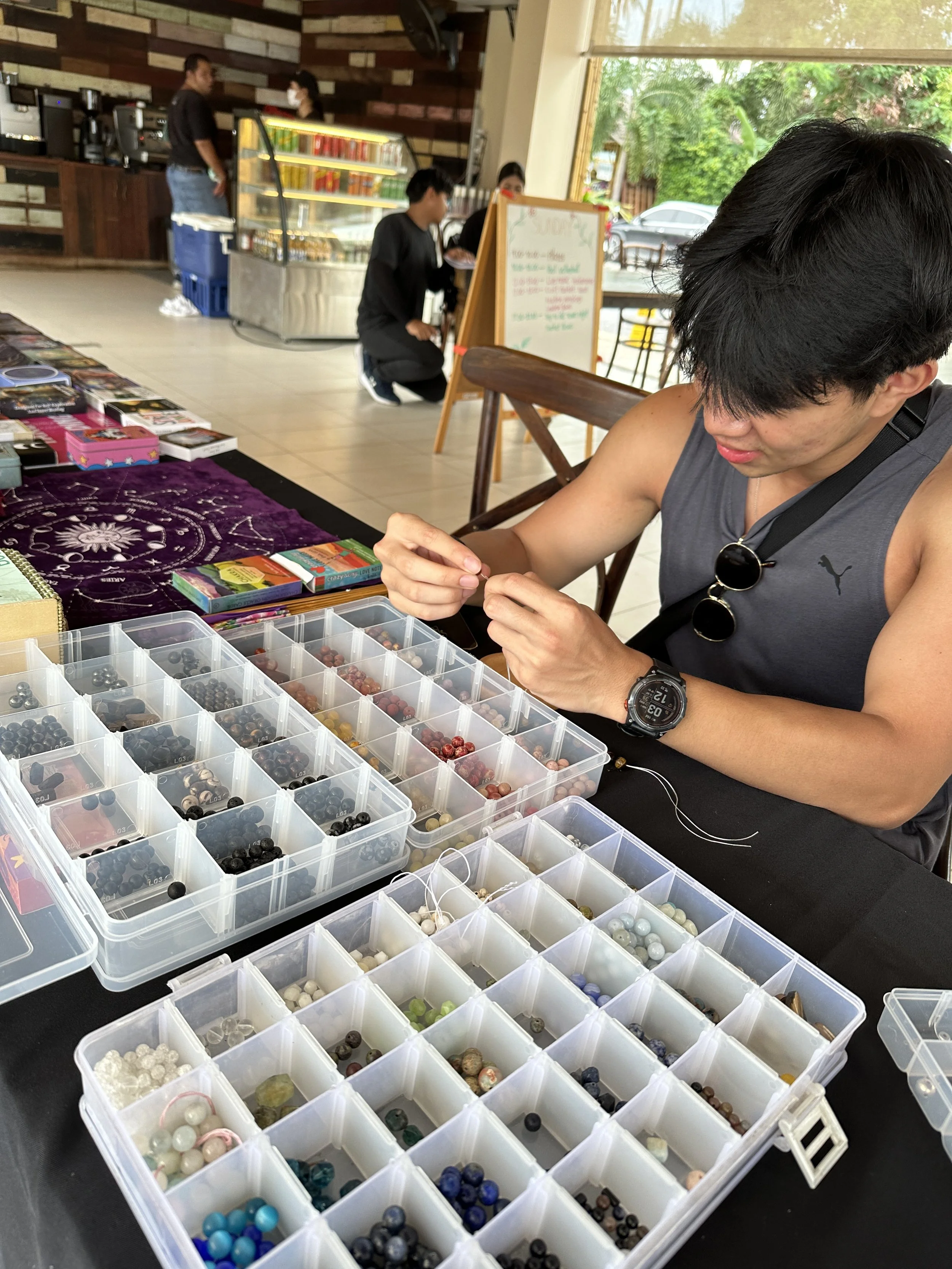 Man with black hair, sleeveless shirt, and sunglasses hanging on shirt seated at a table with container trays filled with various beads, some work in progress with string. In the background, a person kneels near a whiteboard, and two people stand near a glass display case inside a café with large windows and trees outside.