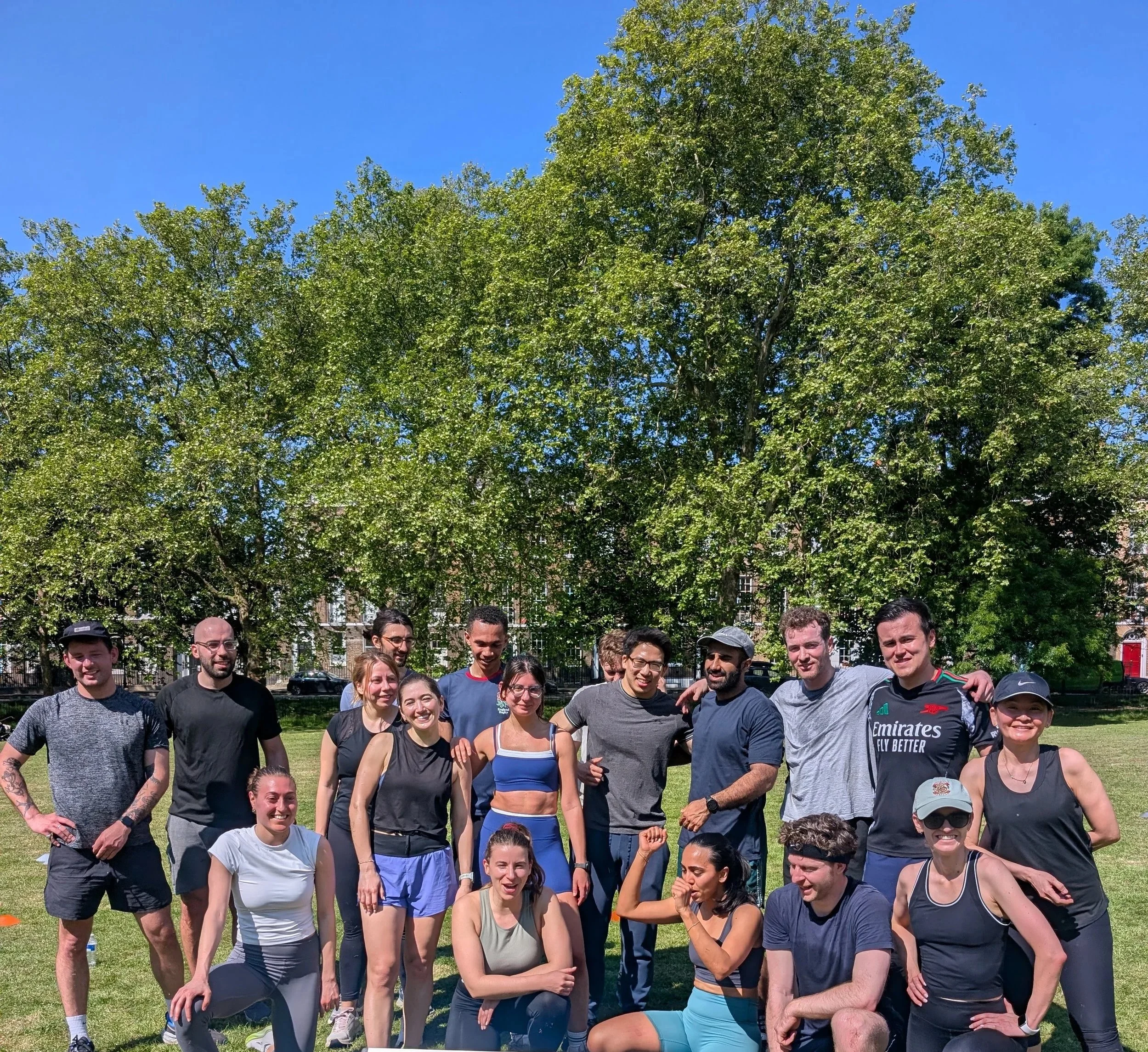 post workout from wild LDN in Highbury Fields, Islington - a group of athletic people sweaty and very happy after the outdoor fitness group class in a beautiful London park on a bright, sunny day, with green trees and a blue sky in the background.