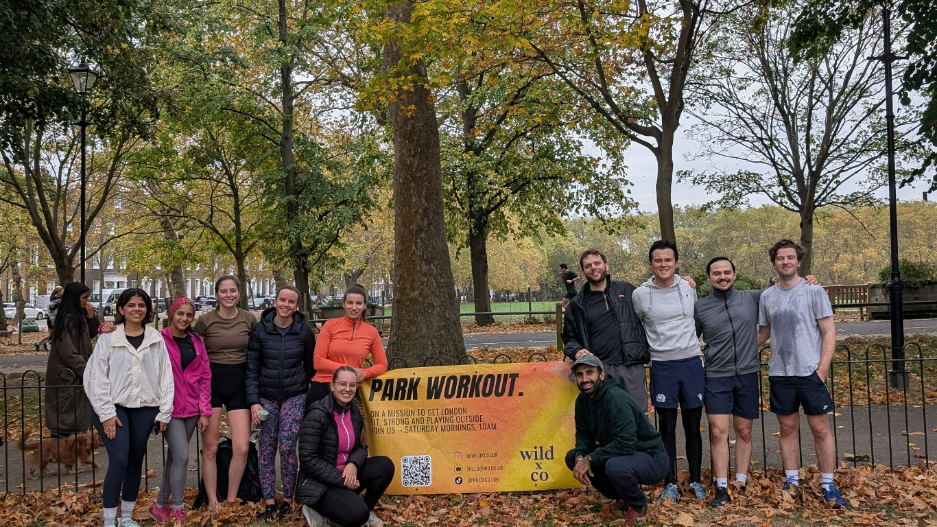 team picture from after a wild LDN session in Highbury Fields, Islington - a group of young, athletic people in their 20s and 30s sweaty and very happy after the outdoor fitness group class in a beautiful park. Autumnal vibes. the sign: PARK WORKOUT.