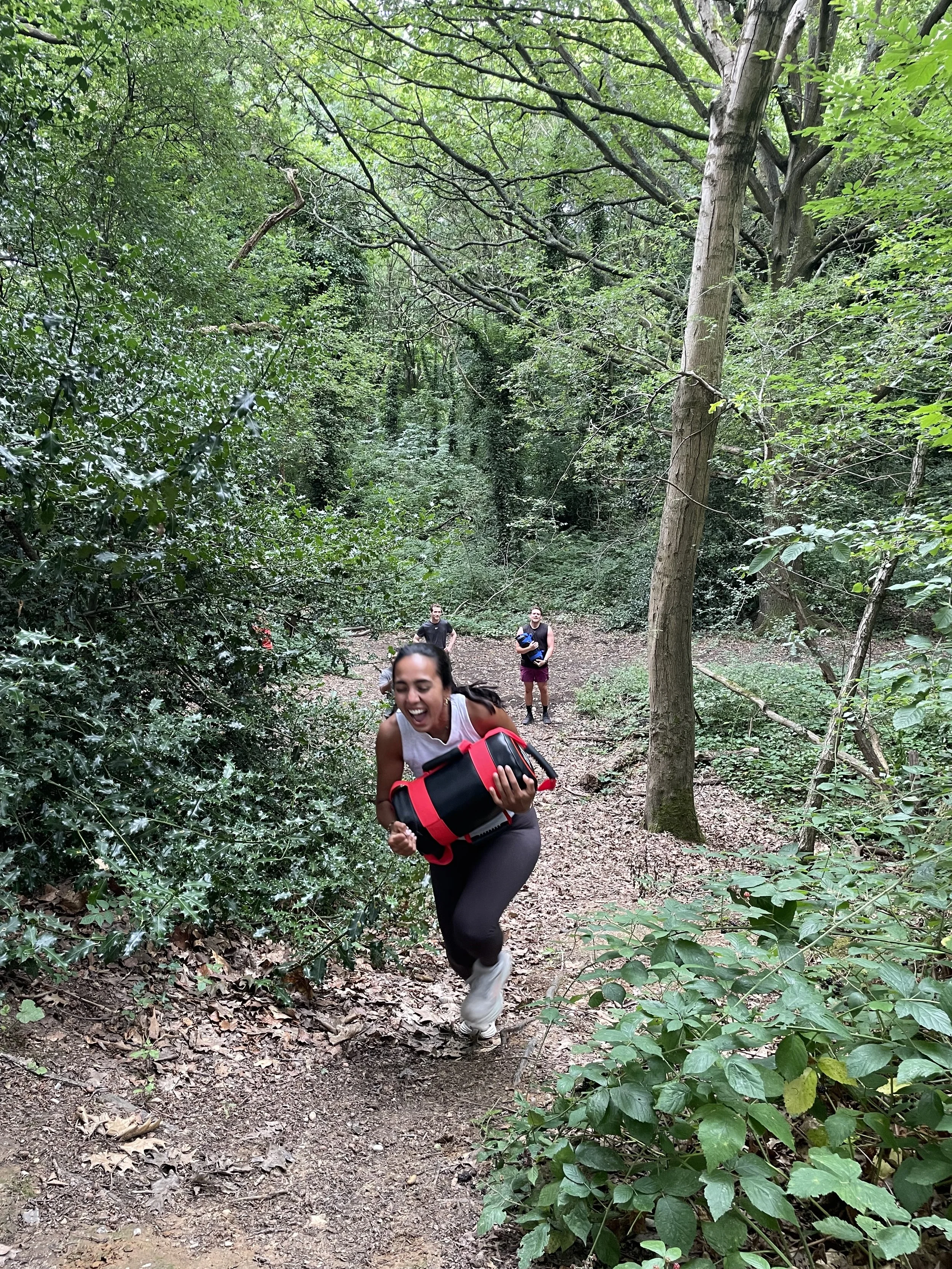 action picture during a wild LDN outdoor fitness class in Hampstead Heath, London where a beautiful woman in her late 20s runs up a hill with a sandbag having lots of fun and the rest of the group watch on