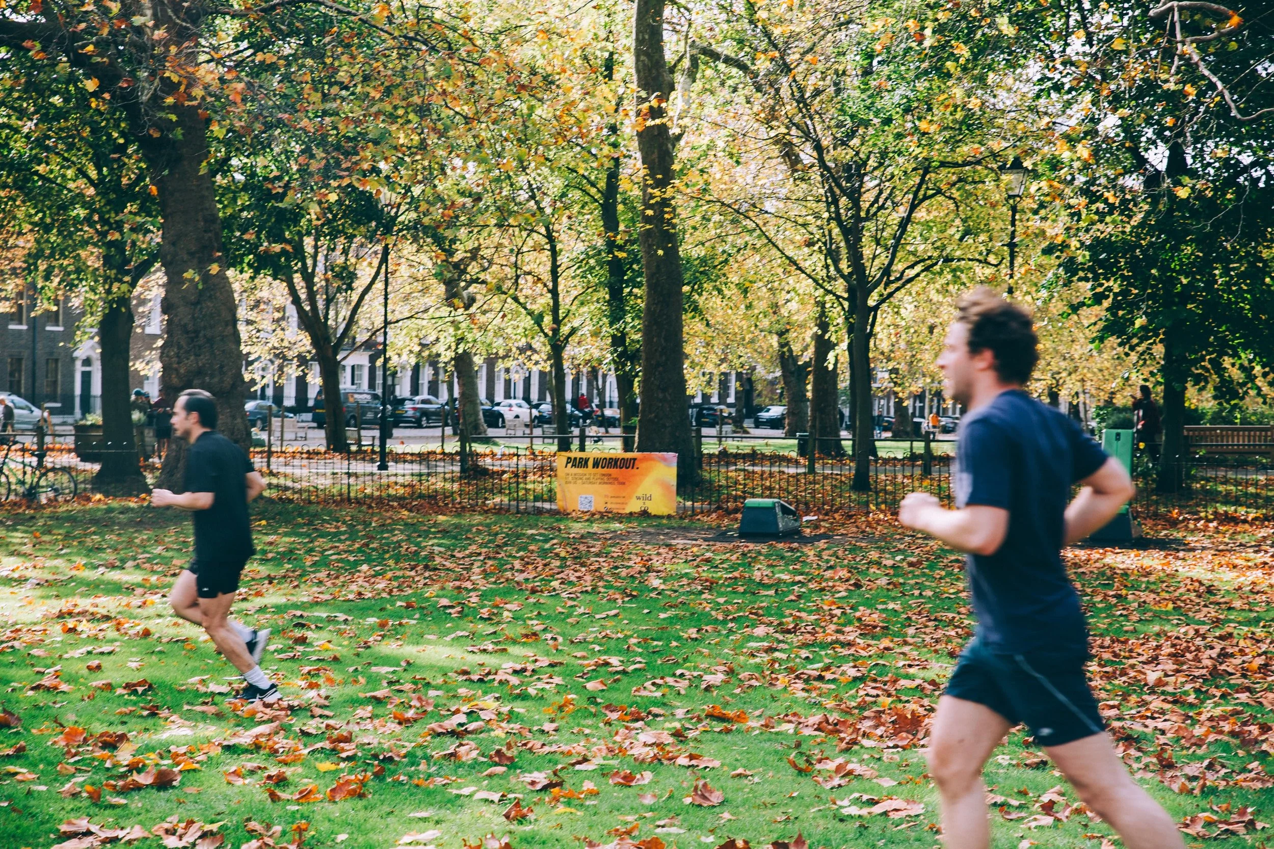 action shot during a wild LDN session in Highbury Fields, Islington - 2 athletic men in their late 20s/early 30s running in the park during the outdoor fitness group class workout. the sign says PARK WORKOUT