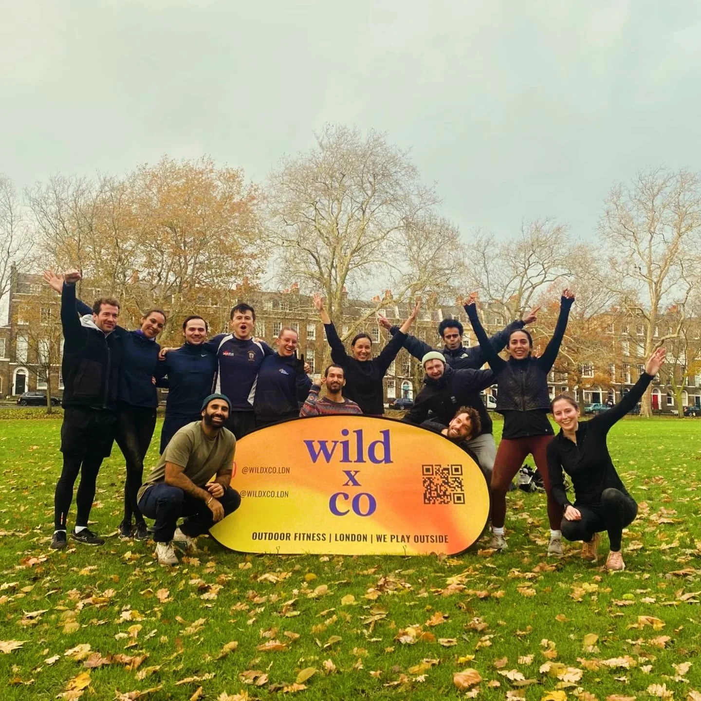  the wild LDN crew happy and cheering after a great outdoor workout class in Highbury Fields, Islington. yellow banner says "wild" with a QR code, social media handles, and the phrases "outdoor fitness," "London," and "we play outside." 