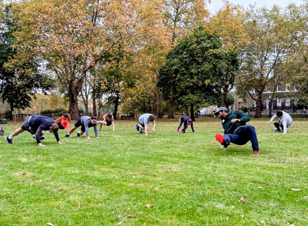 action shot during a wild LDN outdoor workout session in Highbury Fields, Islington - a group of young, athletic people in their 20s and 30s in a circle performing bodyweight movements during the outdoor fitness group class in a beautiful park.