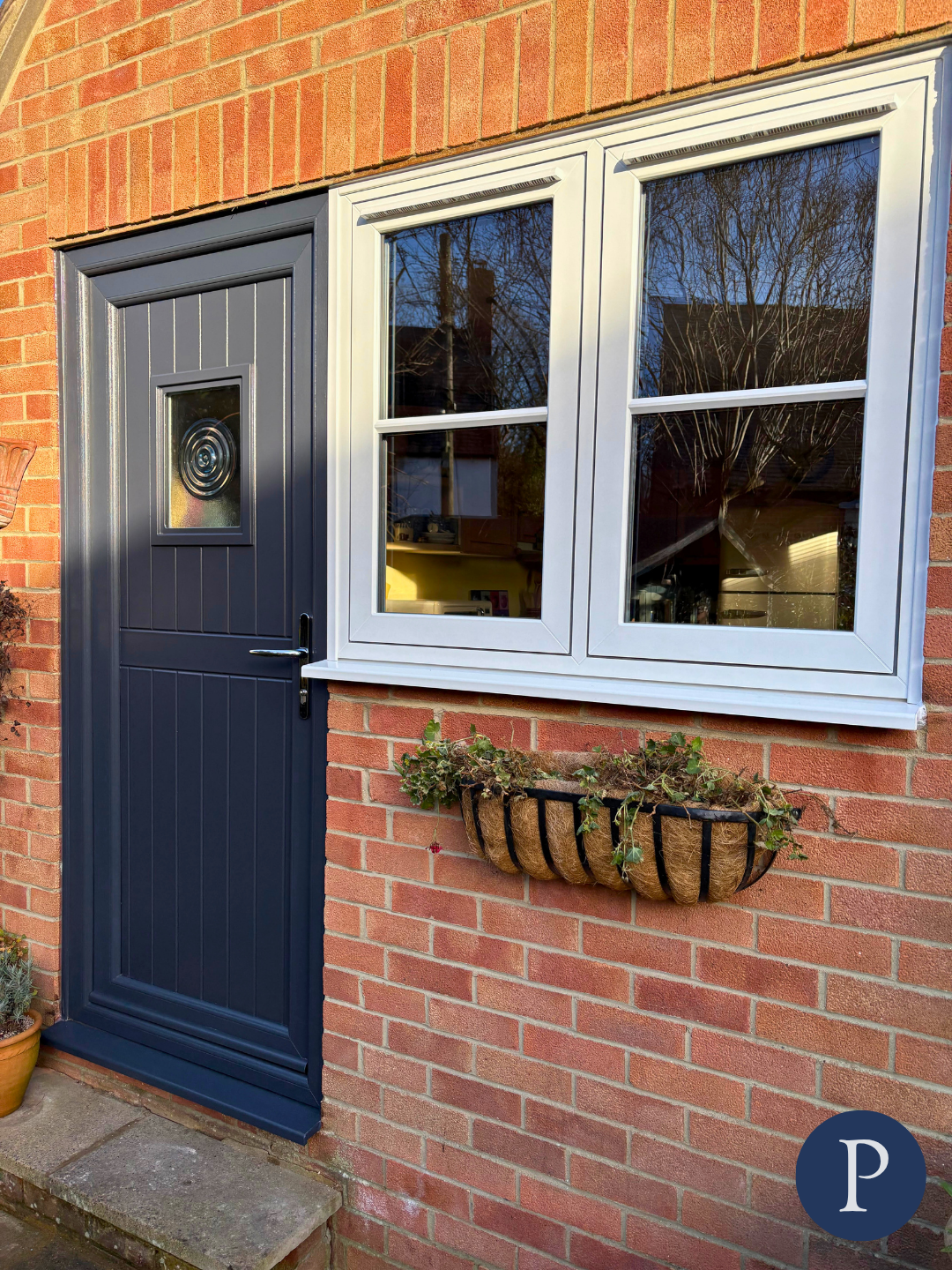 A brick house exterior with a navy blue front door featuring a small circular window and a handle, next to a white-framed window with reflection of a tree and the sky, and a wall-mounted flower basket with green plants beneath the window.