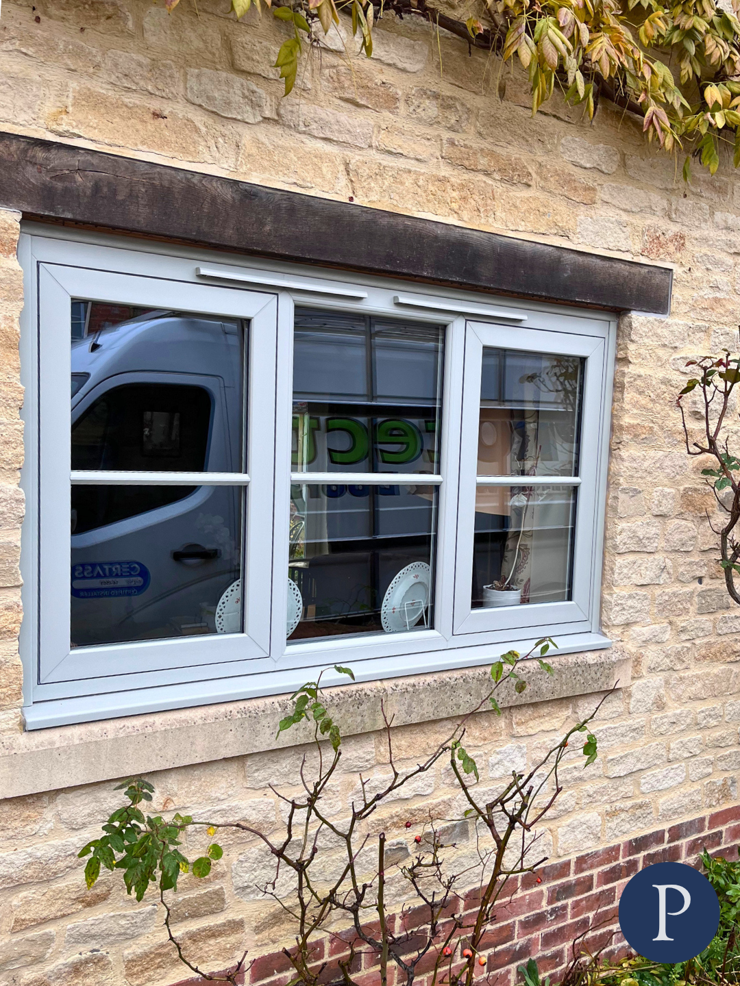 A window with white frames on a brick wall, with a reflection of a gray van and some inside items visible through the glass