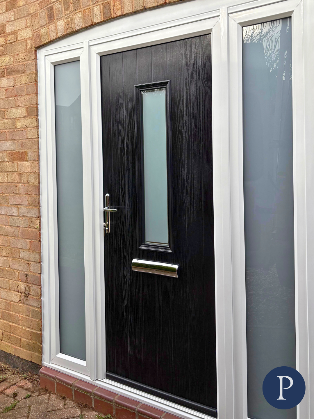 Black front door with a vertical frosted glass window in the middle, a door handle, and a letterbox, surrounded by white frame and flanked by two frosted glass side panels, set in a brick wall.