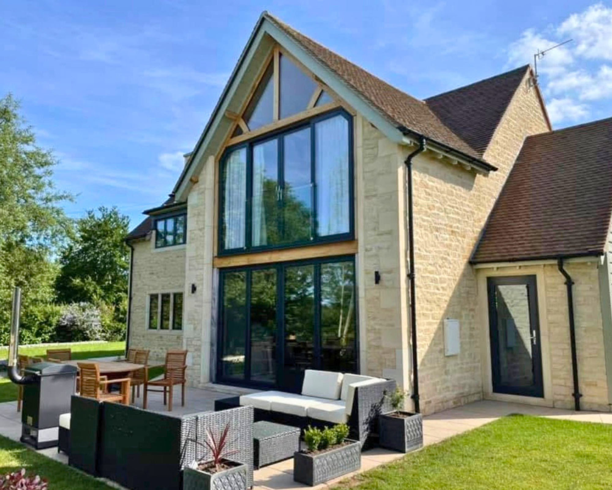 Modern house with large bay window and outdoor patio with seating area surrounded by green lawn and trees.
