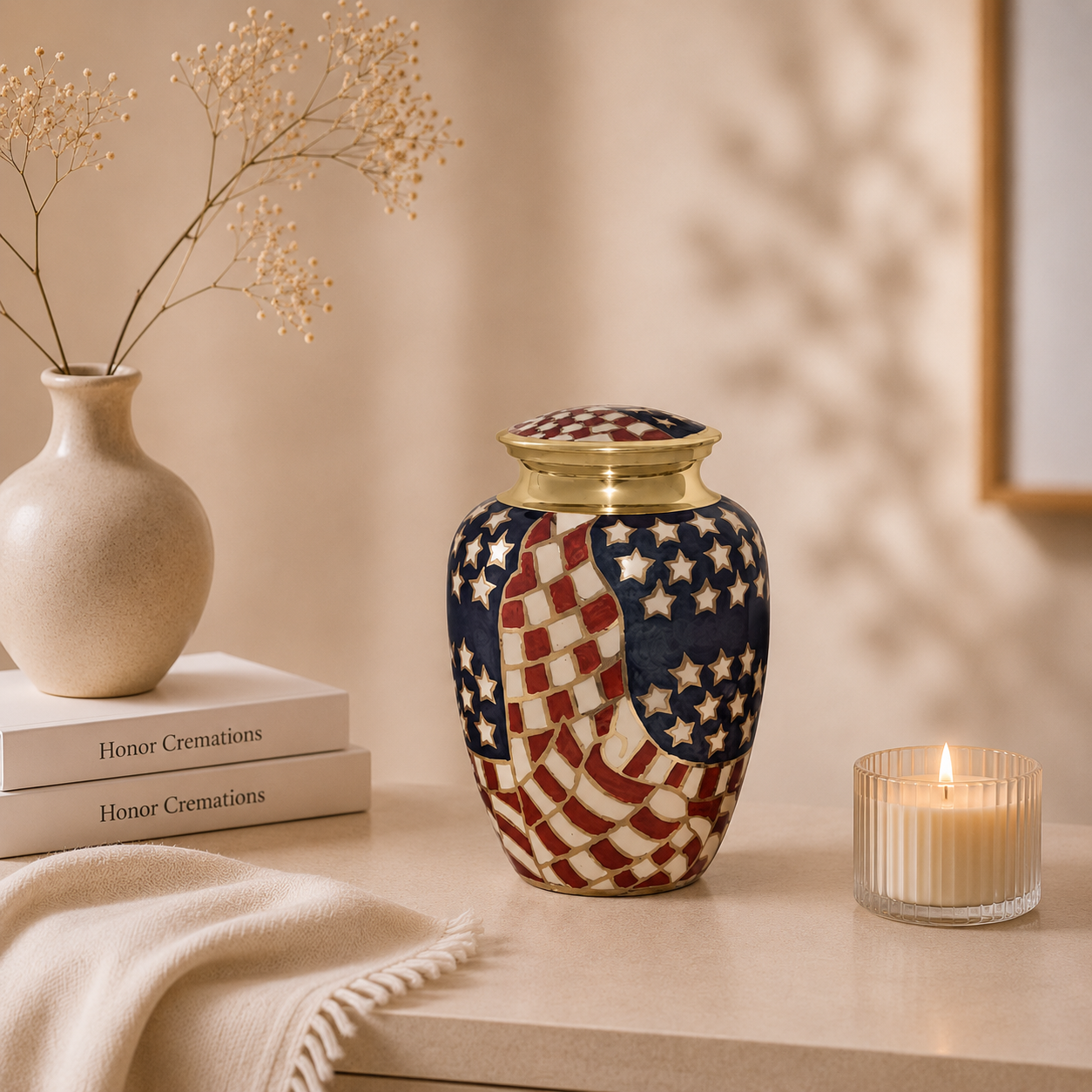 Decorative patriotic-themed urn on a beige surface, with a lit candle, white flowers in a beige vase, and two books titled 'Honor Cremations' in the background.