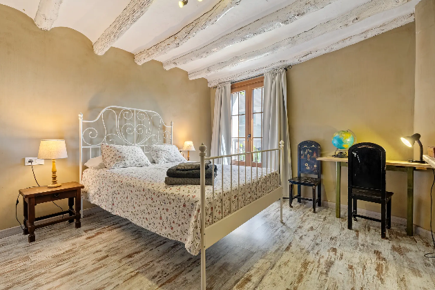 Habitación con cama de metal, dos lámparas, mesita de noche, ventanas con cortinas, escritorio con globo terráqueo y lámpara, y silla.