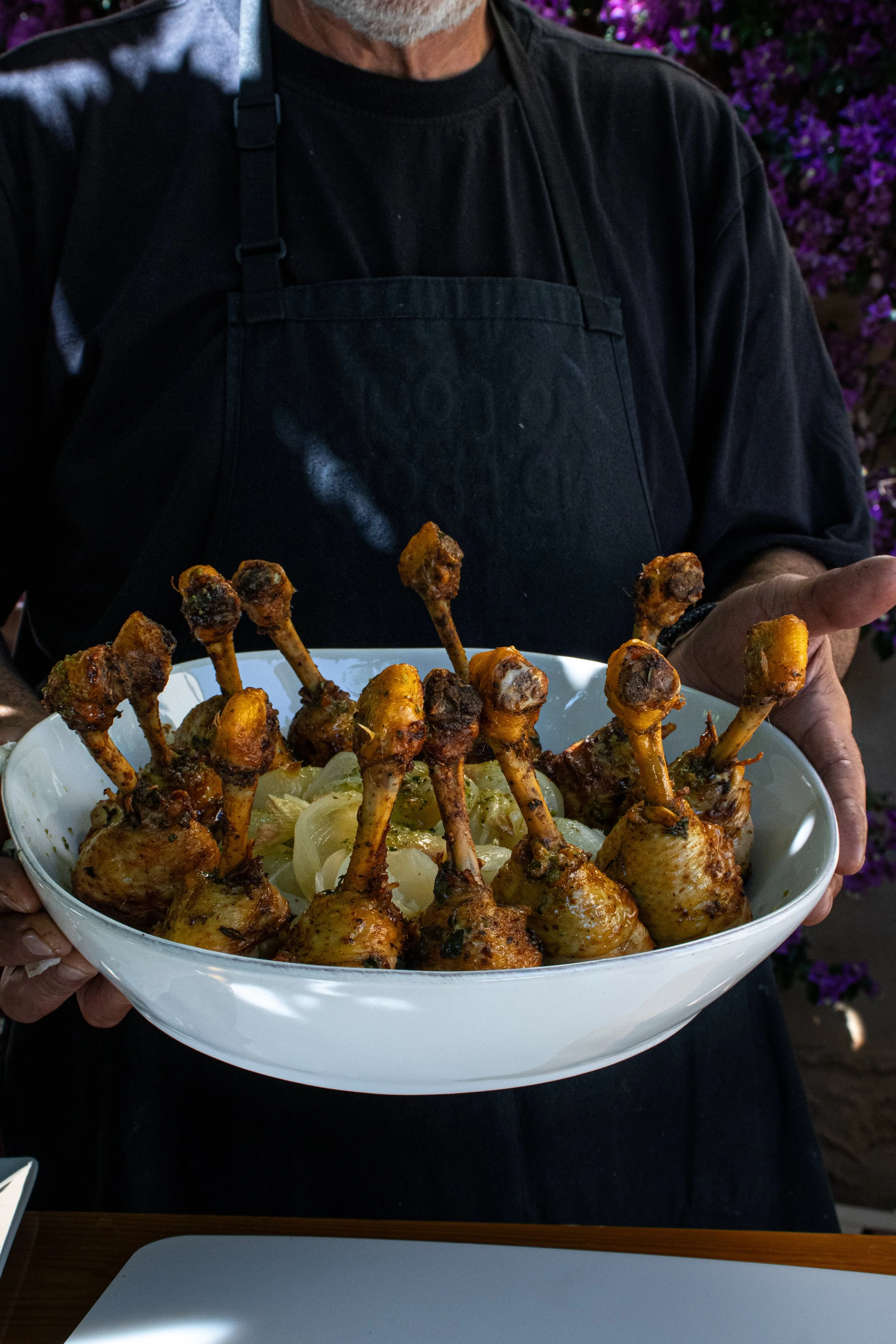 Una persona sostiene un plato grande con muslos de pollo al horno en la parte de arriba, sobre una cama de cebolla y otras verduras, decorado con hierbas.