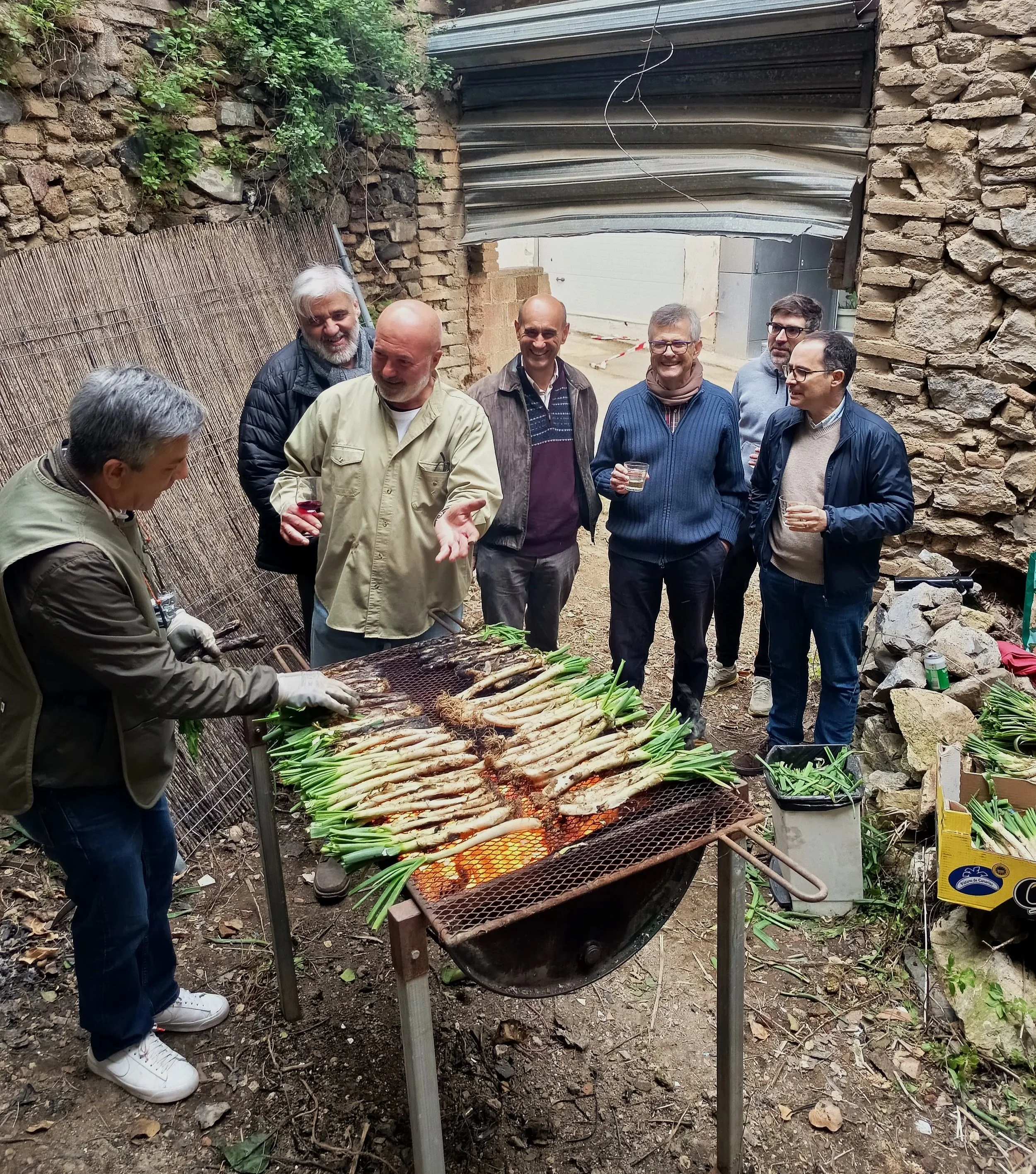 Hombres en una reunión al aire libre, alrededor de una parrilla con verduras asándose, en un ambiente rústico con paredes de piedra y estructuras improvisadas.