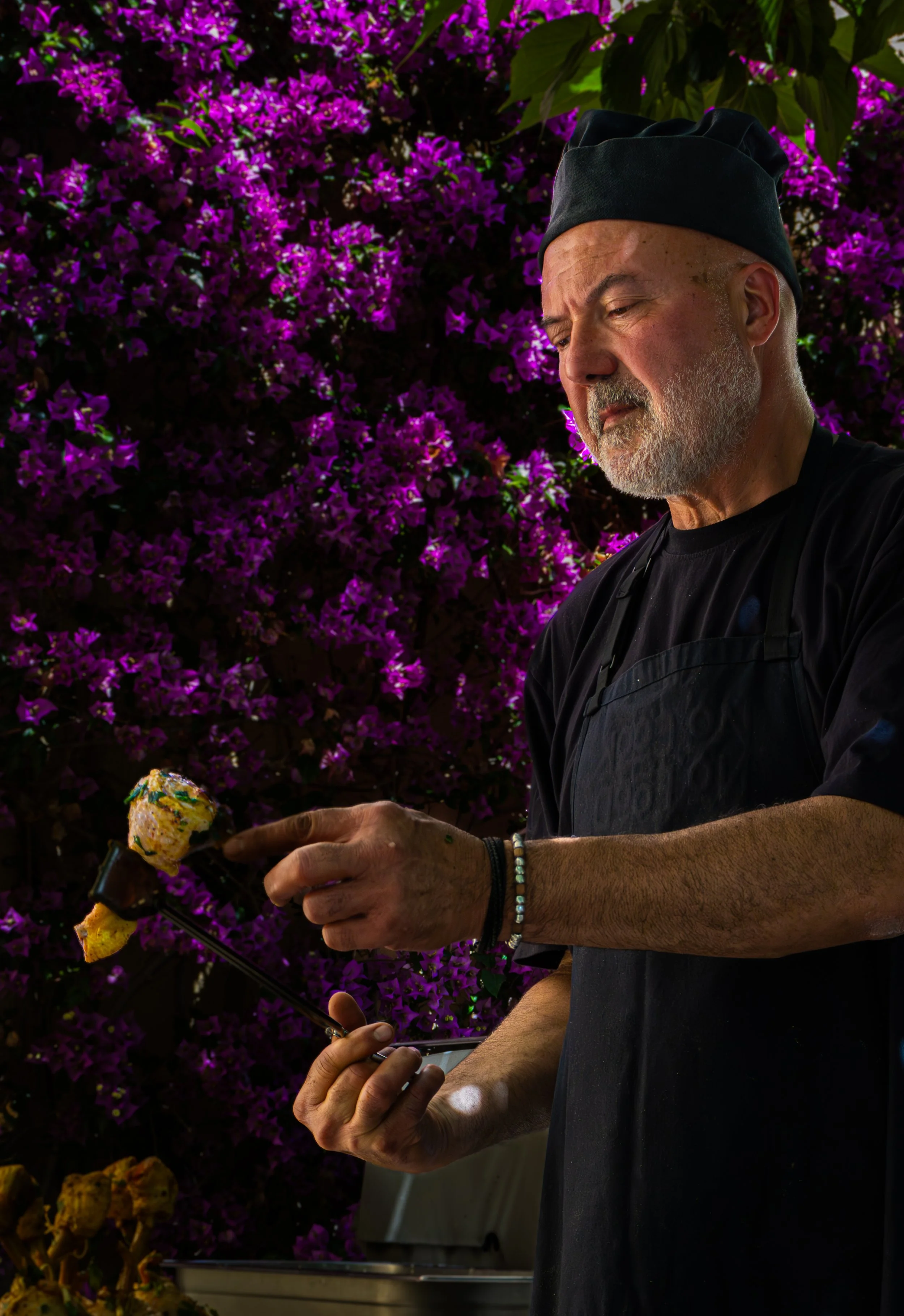 Hombre de cabello canoso y barba, usando un gorro negro y delantal negro, sostiene un trozo de pollo a la parrilla en una mano y un teléfono en la otra, en un fondo de arbustos con flores violetas.