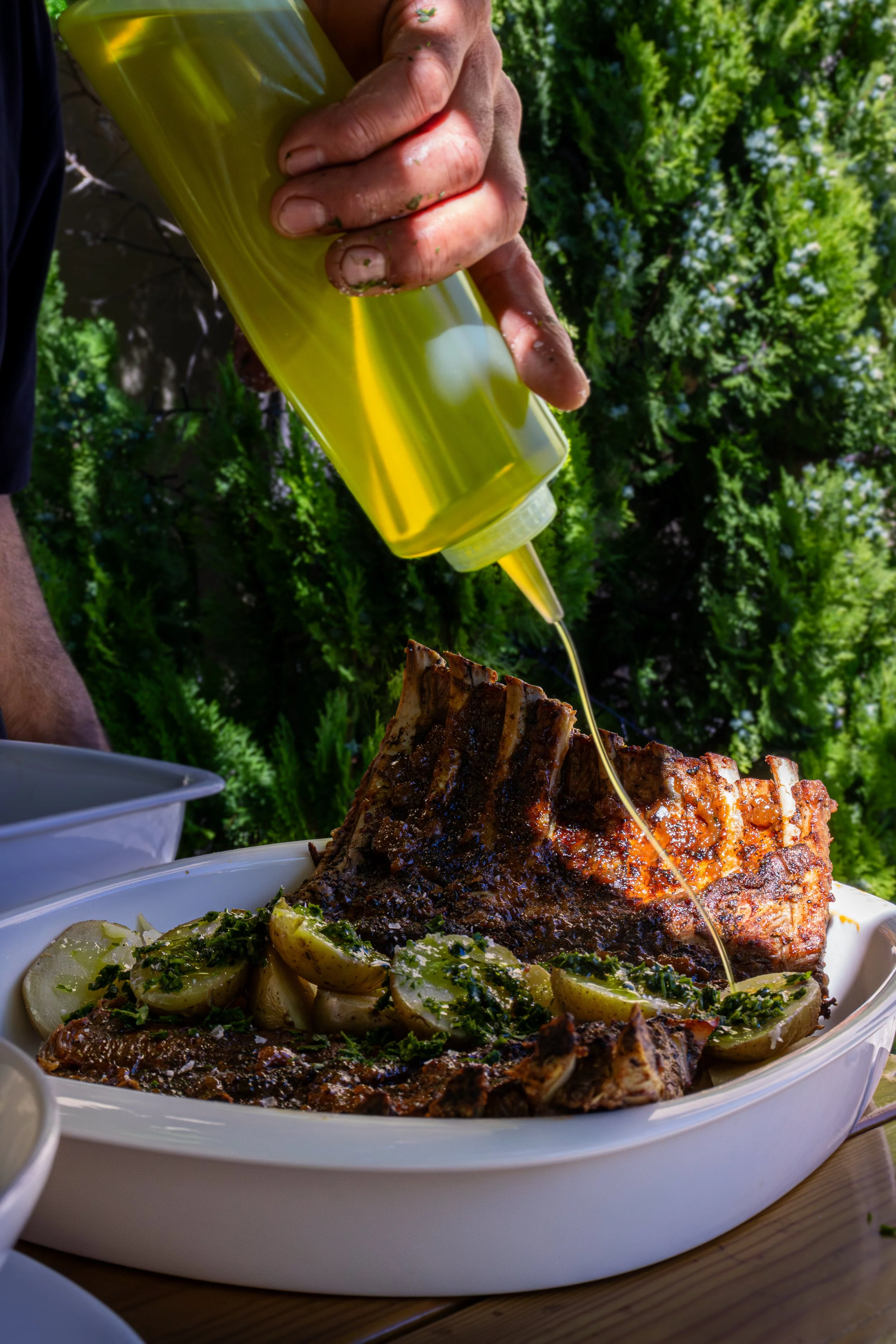 Una comida con costillas de cerdo asadas, acompañadas de patatas y limón, en un plato blanco, con una mano aplicando salsa de mostaza en la carne, en un entorno al aire libre con vegetación de fondo.