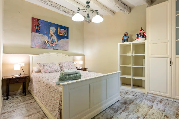 Habitación con cama con ropa de cama floral, dos lámparas de noche, un perchero con cuadros y juguetes en una estantería, y una pared decorada con un cuadro colorido.